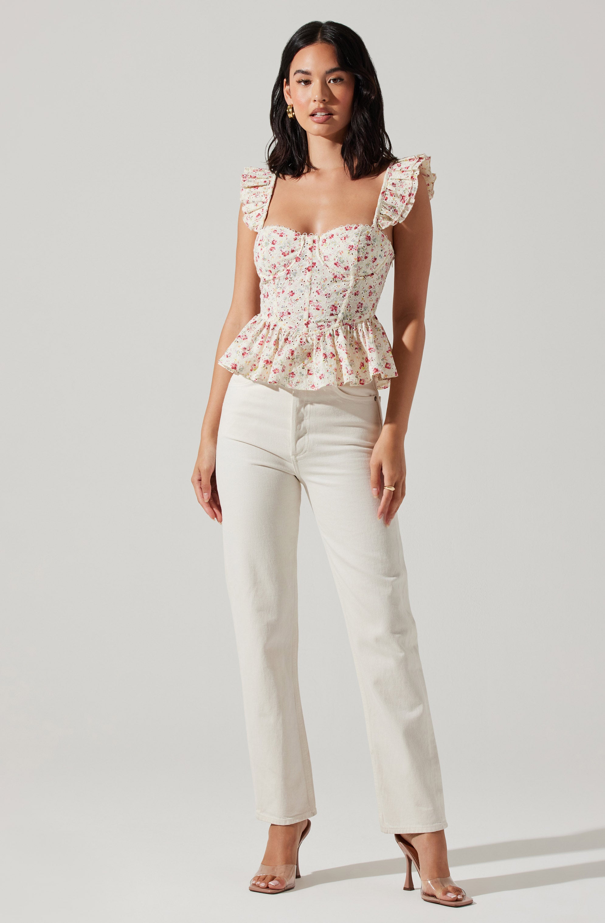 The woman is wearing a sleeveless, fitted top with ruffled shoulder straps and a peplum hem. The top has a light background with a pink and red floral pattern. She is also wearing high-waisted, straight-leg white pants and clear high-heeled shoes.