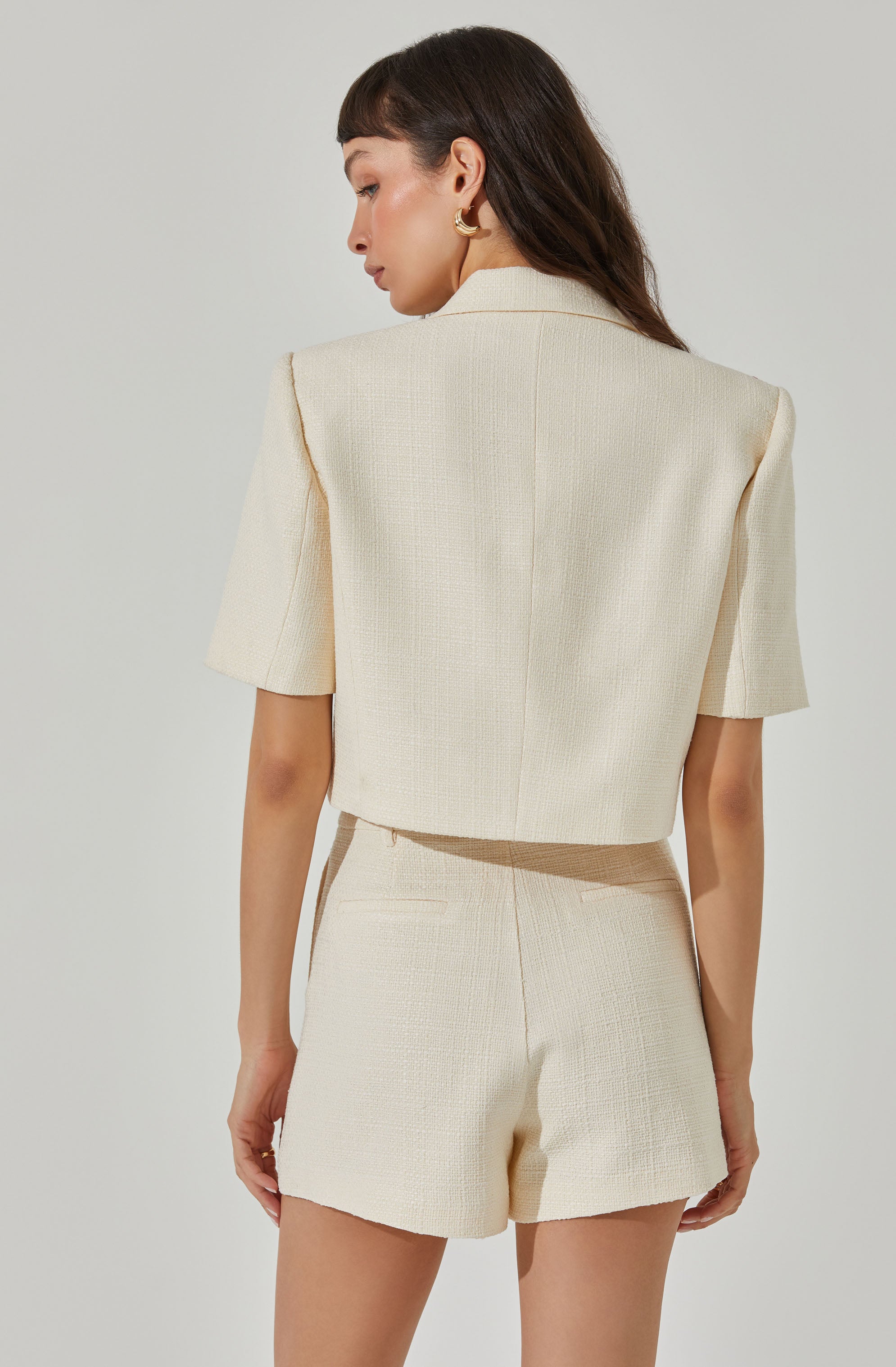A woman is wearing a cream-colored, textured short-sleeve blazer and matching high-waisted shorts. The blazer has structured shoulders and a classic collar, while the shorts have a tailored fit with a single back pocket. She is accessorizing with gold hoop earrings.