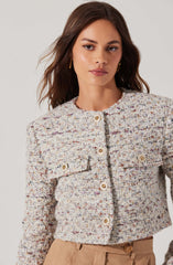A woman is wearing a textured, multicolored, tweed-style jacket with a round neckline. The jacket features decorative gold buttons down the front and on the flap pockets on each side of the chest. She is also wearing tan high-waisted pants.