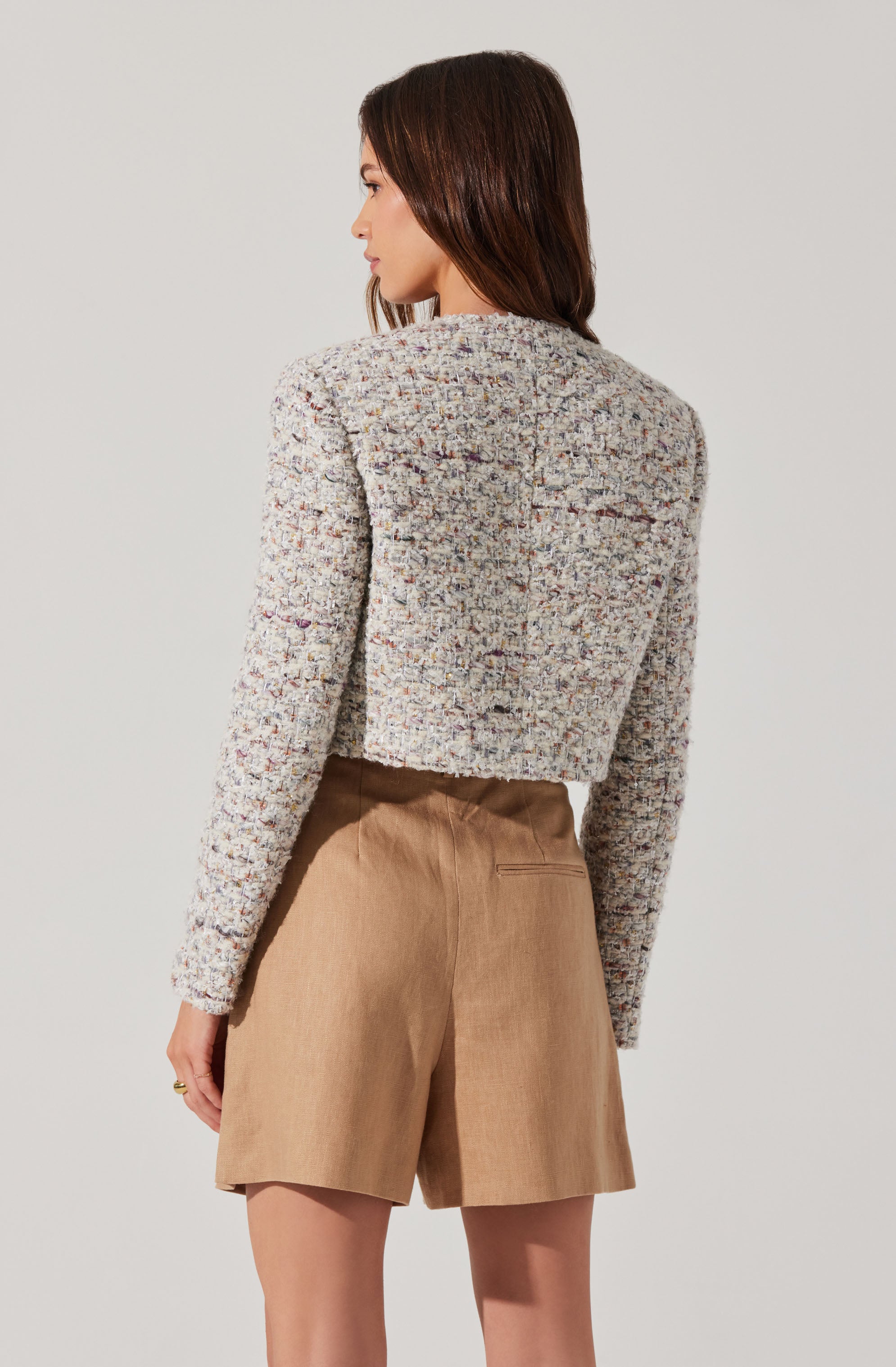 A woman is wearing a textured, multicolored cropped jacket with a boxy fit. The jacket appears to be made of a tweed-like fabric featuring a mix of various colors. She is also wearing high-waisted, light brown shorts with a tailored fit and a back pocket visible.