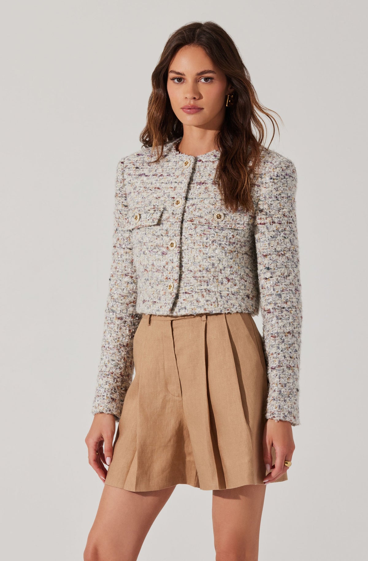 A woman is wearing a textured, multicolored woven cropped jacket with a round neckline and long sleeves. The jacket has decorative gold buttons and a front pocket on the left side. She pairs it with a beige, knee-length pleated skirt made of a lightweight fabric.