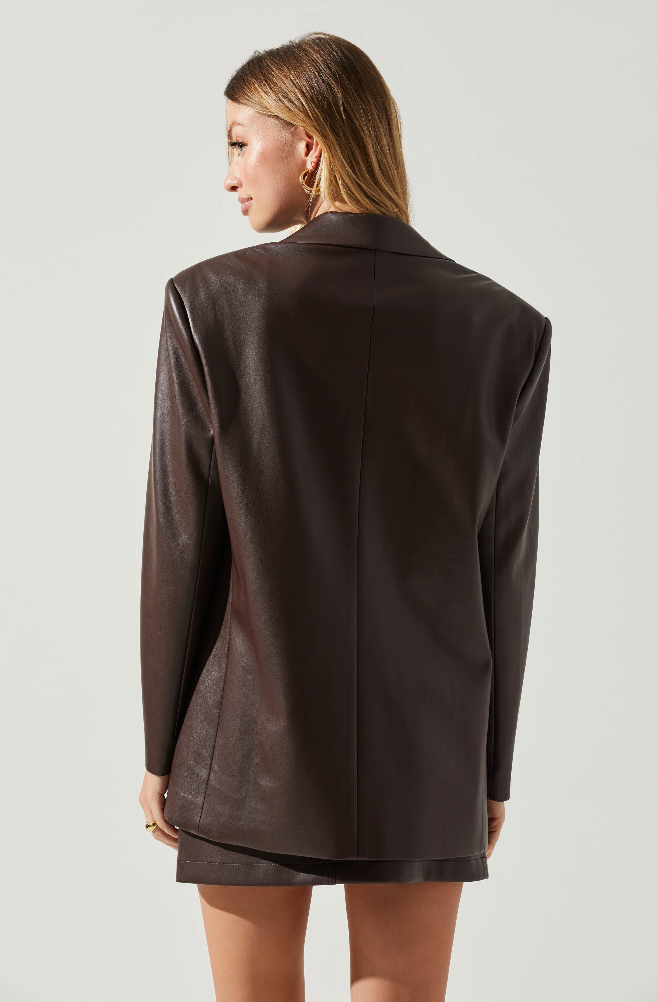 A woman is wearing a dark brown leather blazer with a smooth finish. The blazer has a classic collar and long sleeves, and it extends past her hips. She is also wearing a matching dark brown leather skirt underneath. She has blonde hair and is accessorized with gold hoop earrings and a ring on her right hand.