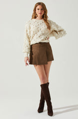 A woman is wearing a cream-colored, chunky knit sweater with a textured, bobble pattern and long sleeves. She pairs it with a high-waisted, brown plaid mini skirt and tall, dark brown suede knee-high boots. She accessorizes with gold and pearl drop earrings and gold rings.