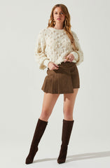 A woman is wearing a cream-colored chunky knit sweater with a textured pattern, paired with a high-waisted brown plaid skirt with pleats. She has on tall, dark brown suede boots that reach just below the knees and is accessorized with gold earrings and rings.