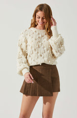 A woman is wearing a chunky cream-colored sweater with a textured pattern featuring large raised loops and small pom-poms, paired with a brown plaid mini skirt. She has long, wavy ginger hair and is accessorized with large, gold, teardrop earrings and rings on her fingers.