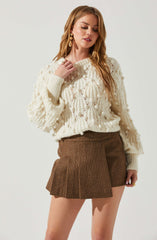 A woman is wearing a cream-colored, chunky knit sweater with a textured, pom-pom detail throughout. She pairs it with a high-waisted, brown plaid pleated skirt. She accessorizes with large, gold drop earrings and rings on her fingers.