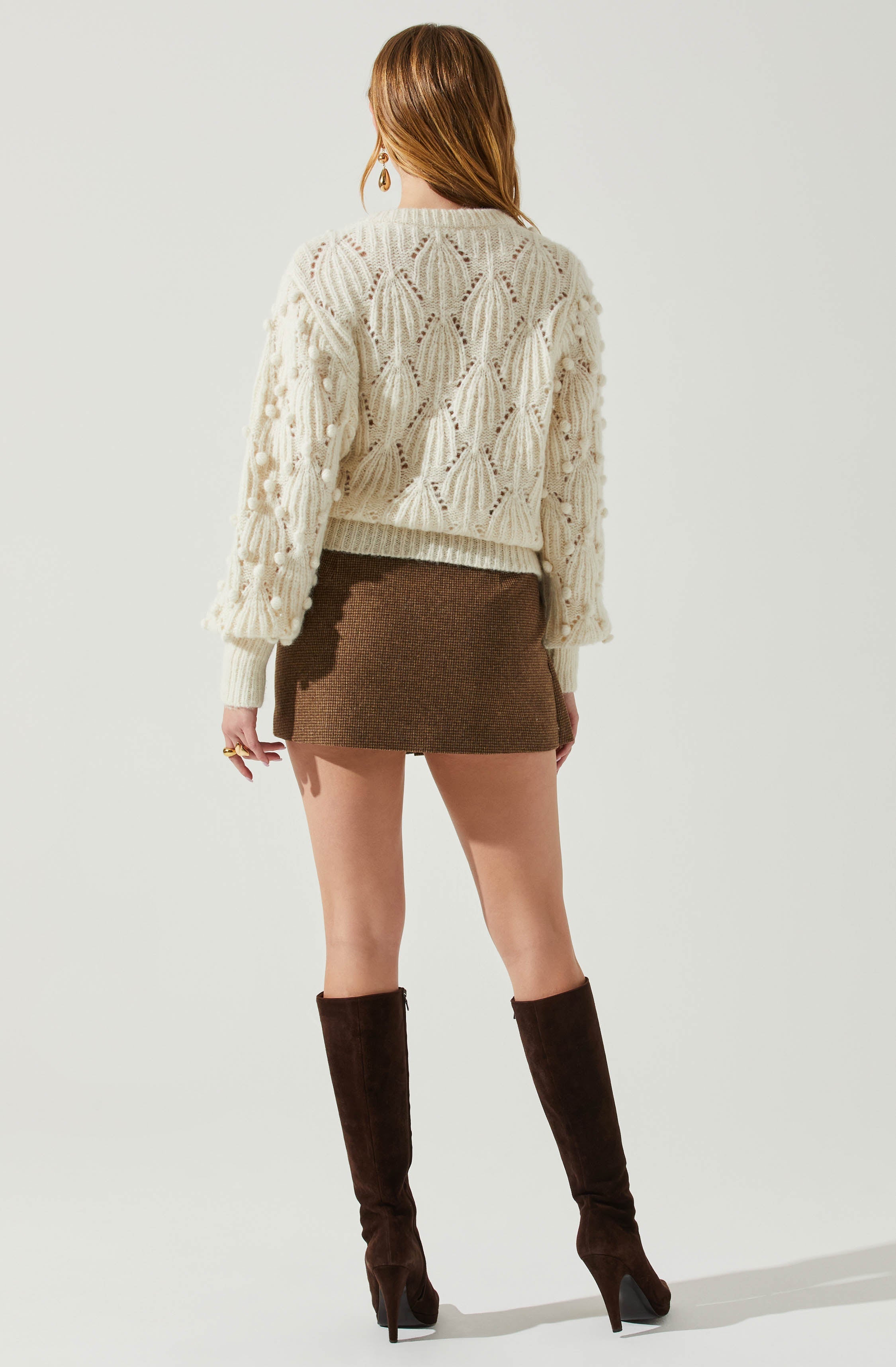 A person is wearing a cream-colored, chunky knit sweater with a textured, openwork pattern and pom-pom details on the sleeves. They are also wearing a short, fitted, brown tweed skirt and knee-high, dark brown suede stiletto boots.