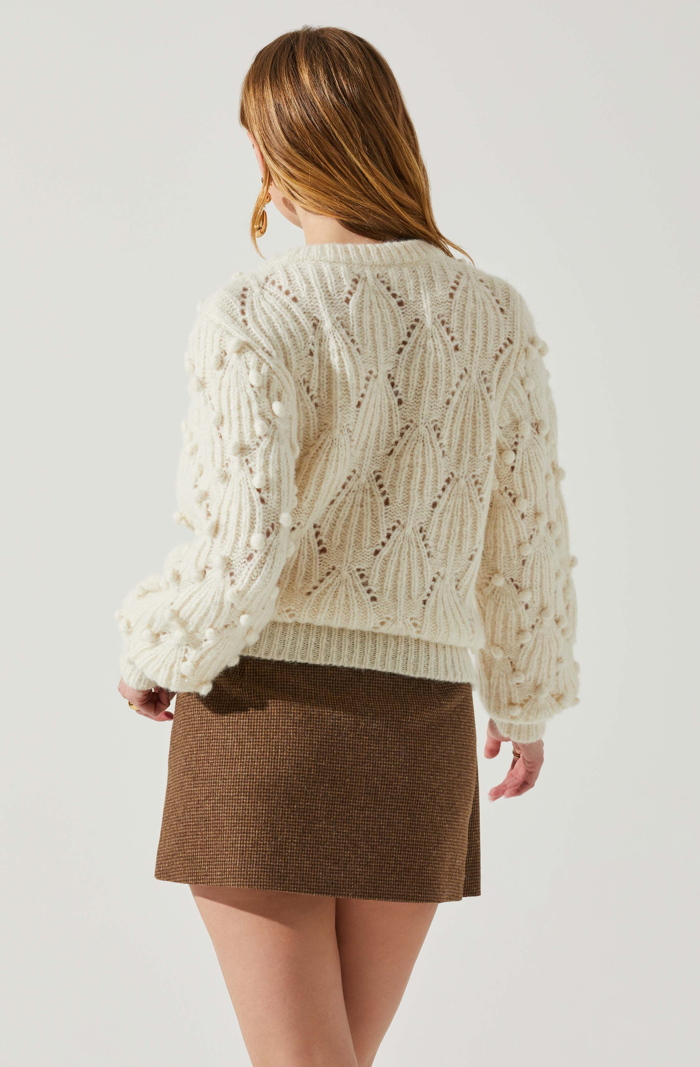 A woman is wearing a cream-colored, chunky knitted sweater with intricate cable and pom-pom details. She pairs it with a high-waisted, brown houndstooth mini skirt.