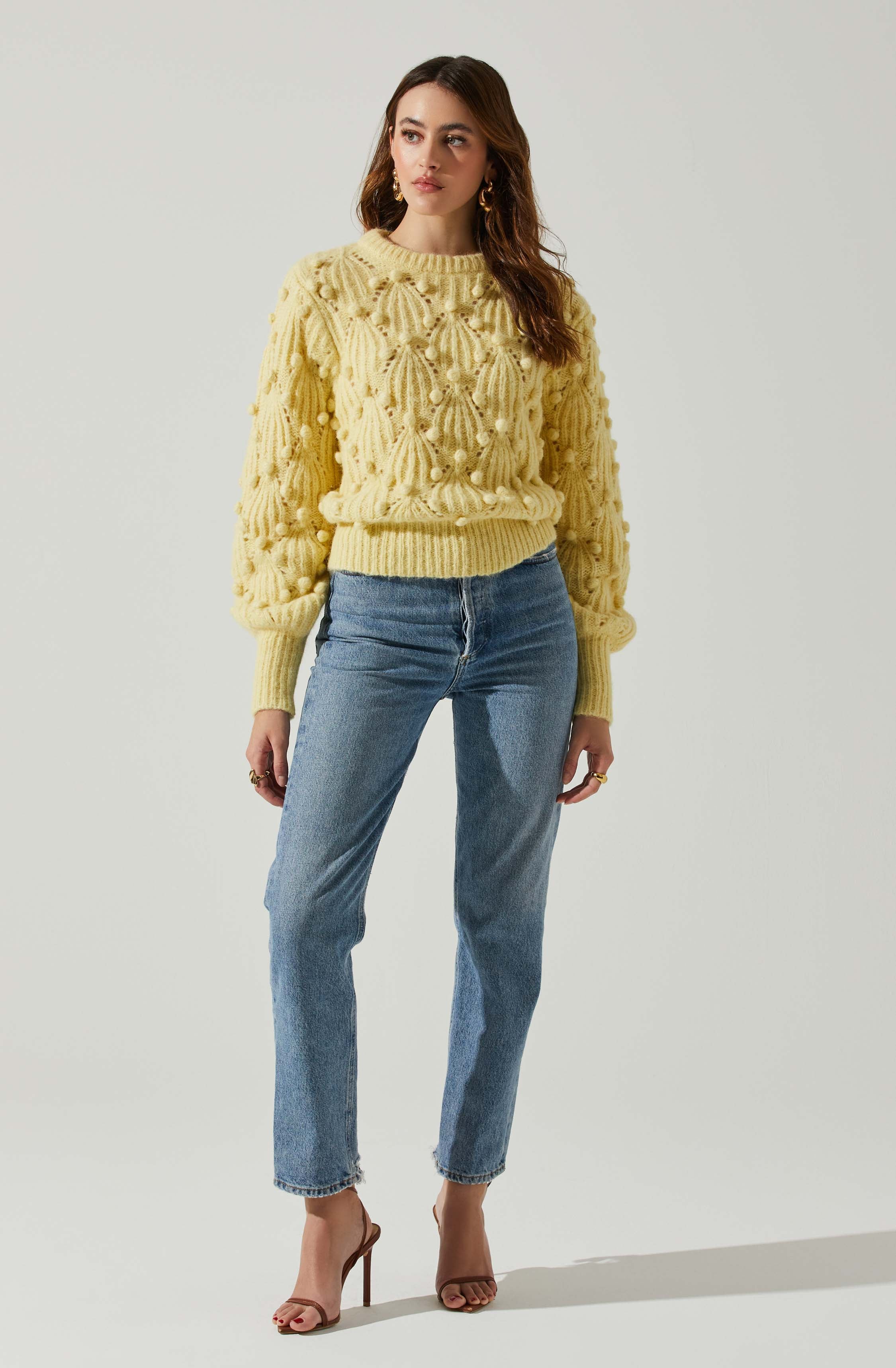 A woman is wearing a chunky, textured yellow sweater with a raised, patterned knit design featuring rounded and ribbed motifs. The sweater has a round neckline, long sleeves, and a ribbed hem. She is also wearing high-waisted, straight-leg blue jeans with a slightly faded wash, paired with open-toe, high-heeled sandals.