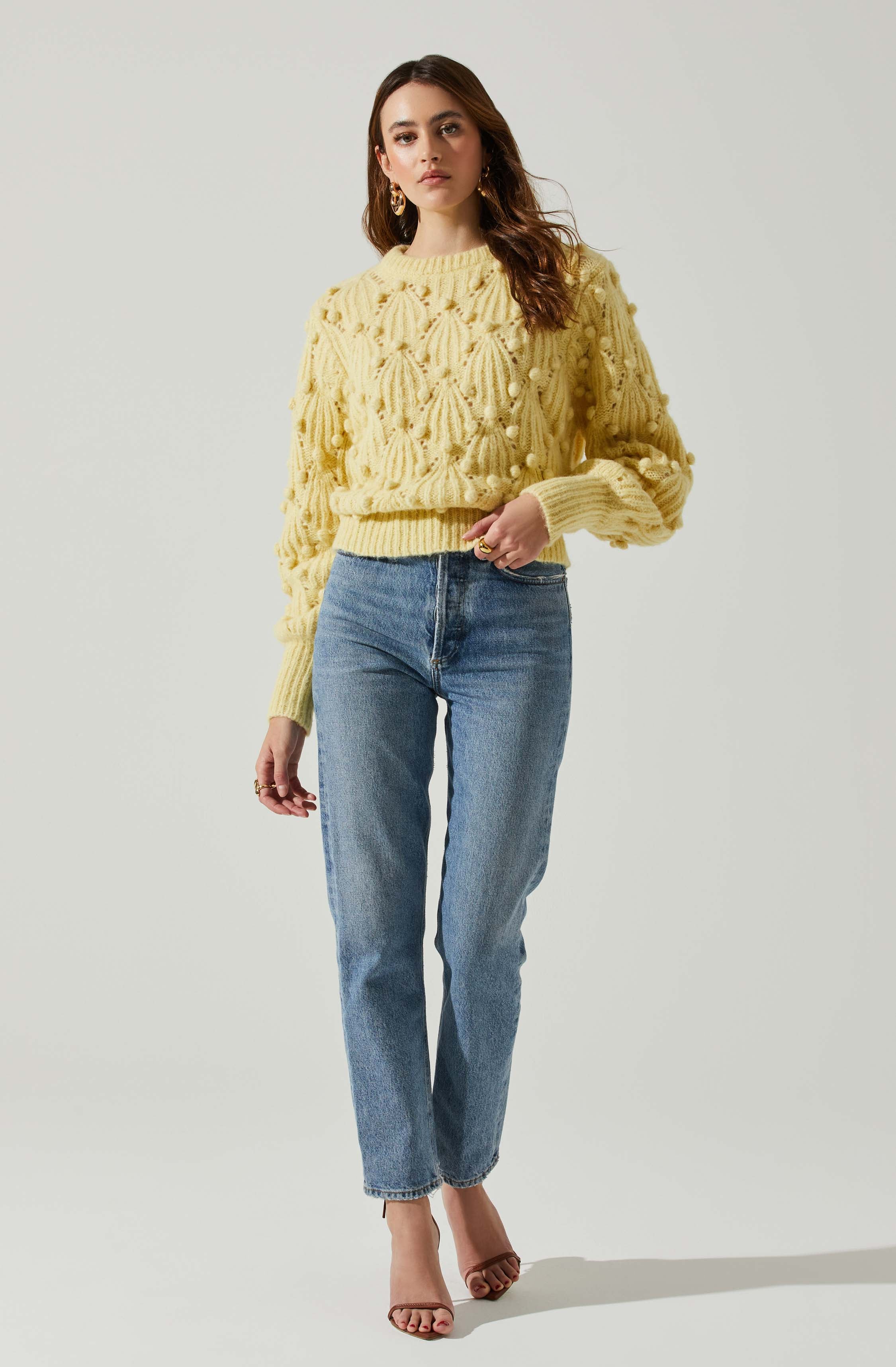 A woman is wearing a yellow chunky knit sweater with a textured pattern and pom-pom accents, paired with high-waisted blue jeans and clear heeled sandals.