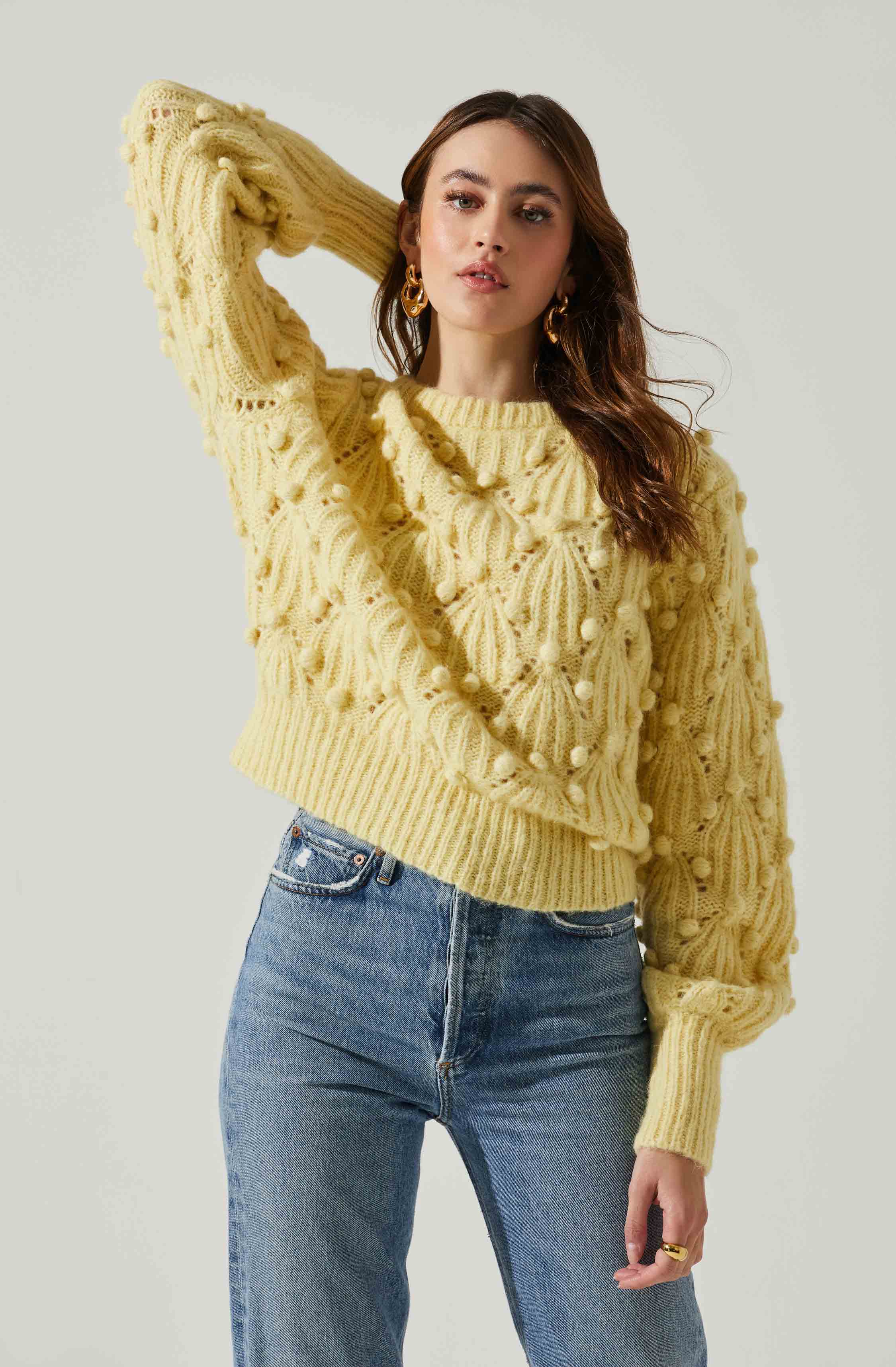 A woman is wearing a chunky, oversized yellow cable-knit sweater with textured pom-pom details throughout. She has paired it with high-waisted blue jeans. She accessorizes with large, gold hoop earrings and a gold ring. Her dark brown hair is styled in soft waves.
