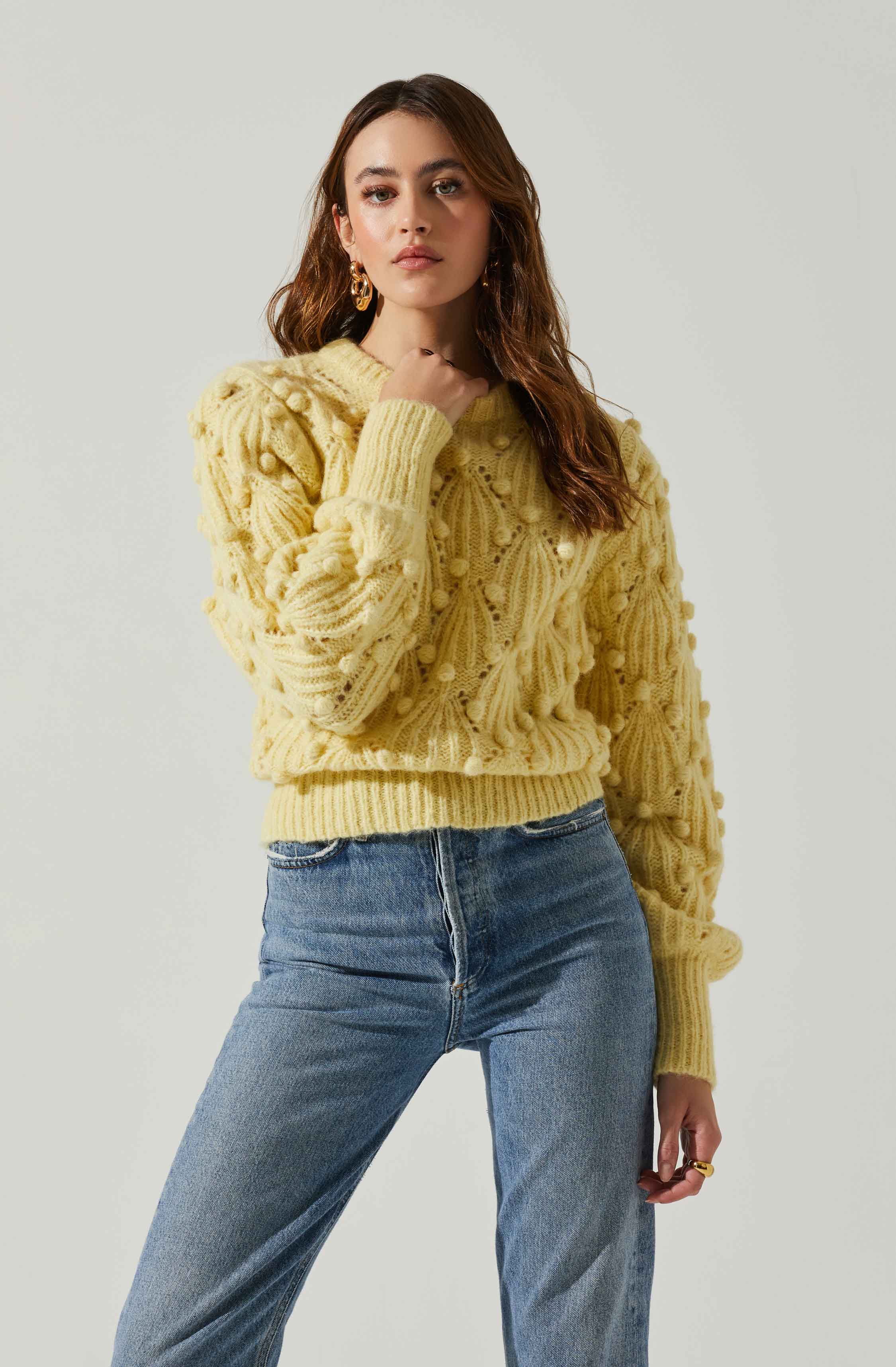 A woman is wearing a yellow, chunky knit sweater with textured, pom-pom-like details. The sweater has a crew neckline and long sleeves. She pairs it with high-waisted blue jeans. She has long, wavy brown hair and is accessorized with gold hoop earrings and a gold ring.
