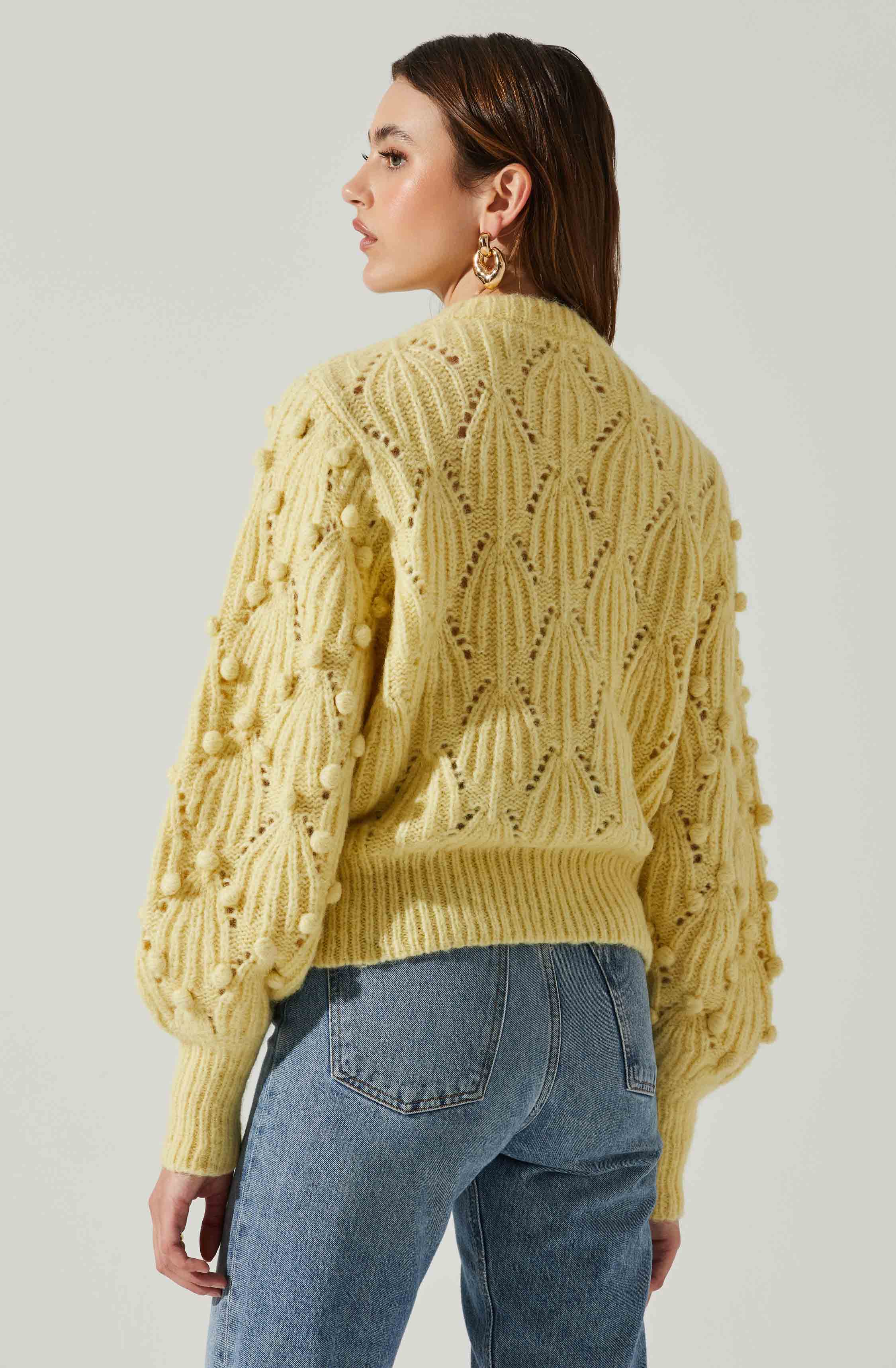 A woman is wearing a chunky, yellow, cable-knit sweater with a pattern of open lacework and pom-pom embellishments on the sleeves. She pairs it with high-waisted blue jeans. She has shoulder-length brown hair and is accessorized with large gold hoop earrings.