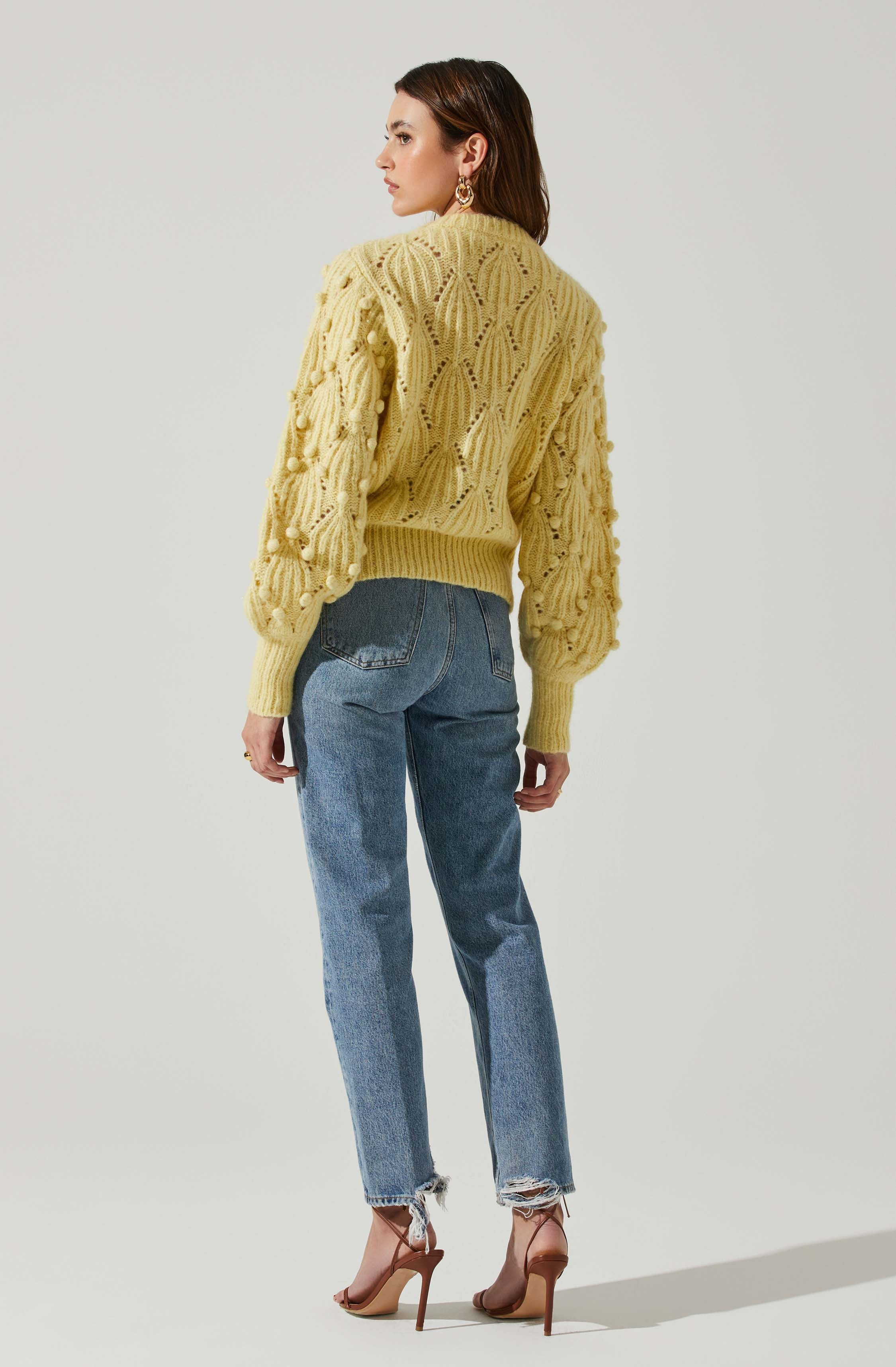 A woman is wearing a yellow chunky knit sweater with a textured, openwork pattern and pom-pom embellishments on the sleeves. She pairs it with high-waisted, slightly distressed blue jeans and nude high-heeled sandals. She also has on large, circular earrings.