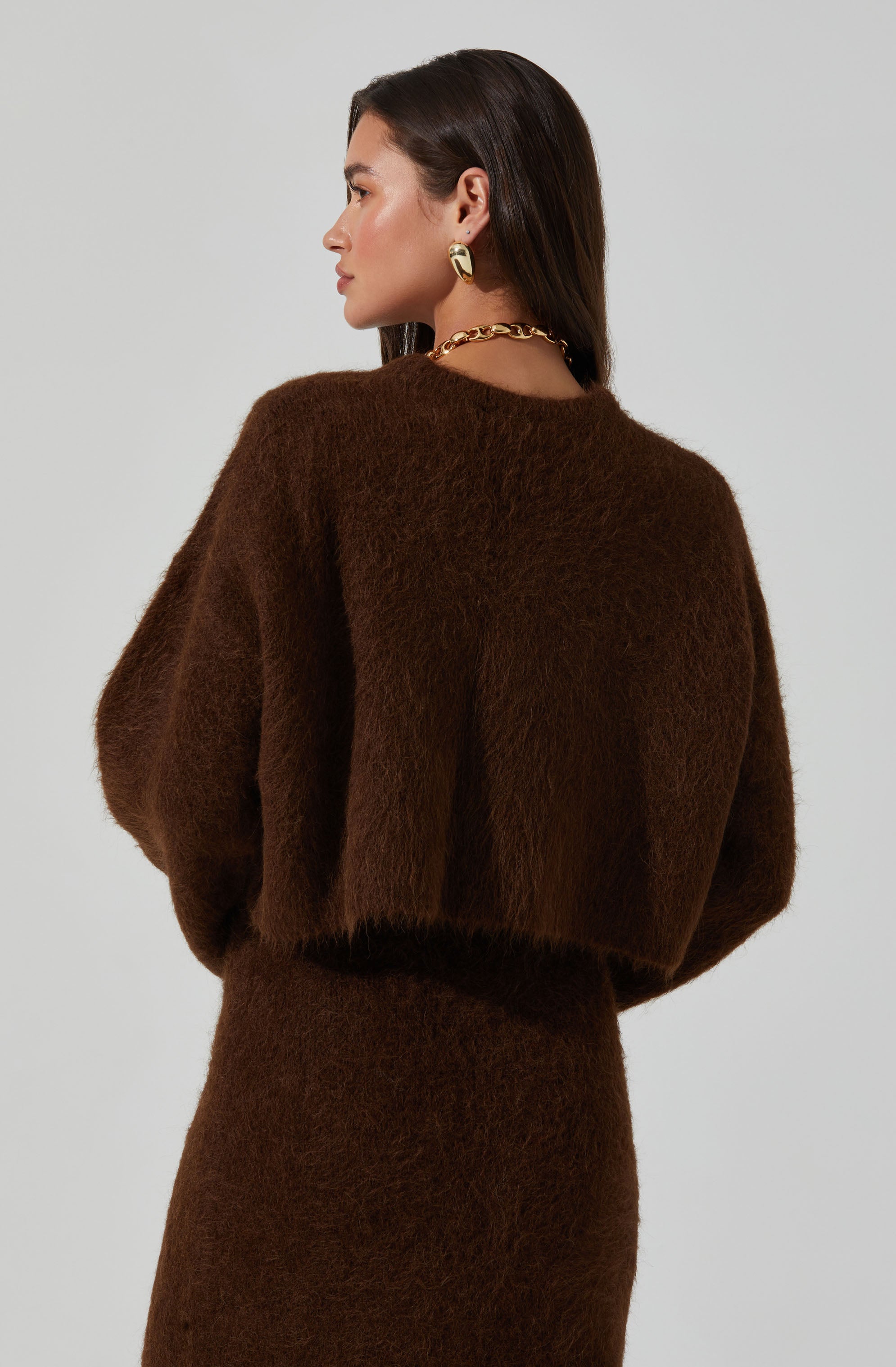 A woman is wearing a dark brown, fuzzy, long-sleeved sweater and a matching skirt. She has accessorized with large gold hoop earrings and a chunky gold chain necklace.