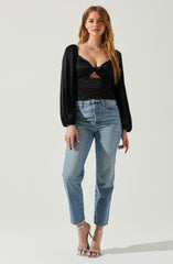 A woman is wearing a black satin top with a twist-front detail and a cutout at the midriff, paired with a black long-sleeve jacket with puffed sleeves. She is also wearing high-waisted, light blue jeans with a slightly relaxed fit, and silver high-heeled sandals with ankle straps and decorative embellishments.