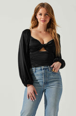 A woman with long, wavy, reddish-brown hair is wearing a black satin long-sleeve top with puffed shoulders, a twisted front detail, and a small cutout below the bust, accented with a rhinestone trim along the neckline. She is also wearing light blue high-waisted jeans with a relaxed fit.
