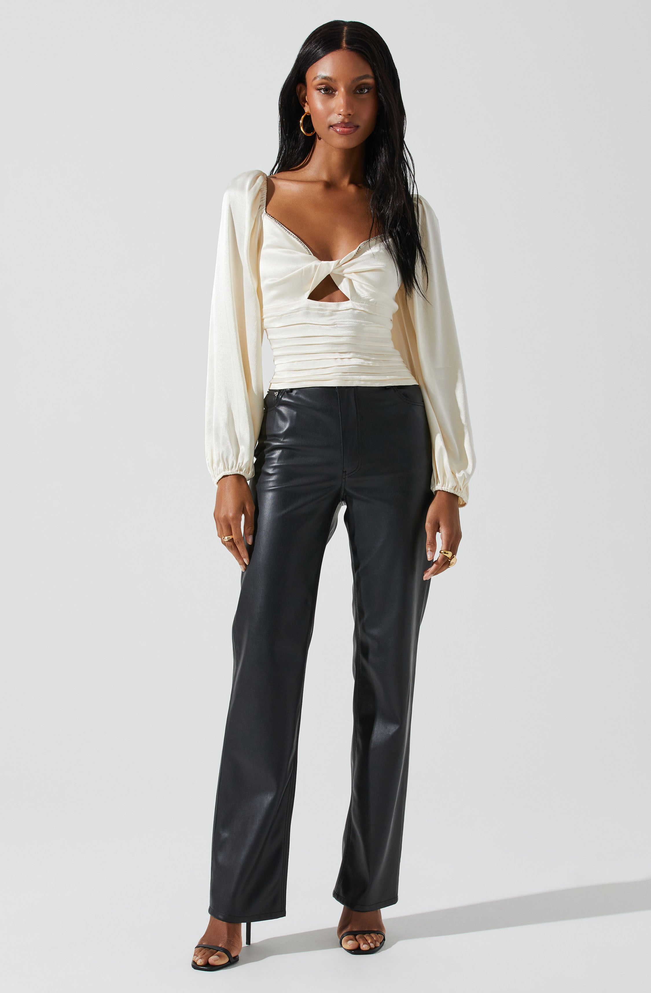A woman is wearing a cream-colored, long-sleeved blouse with a large front knot and a small cutout below the knot. She pairs it with high-waisted, black leather wide-leg pants. She accessorizes with gold hoop earrings and rings, and is wearing black open-toe high heels.