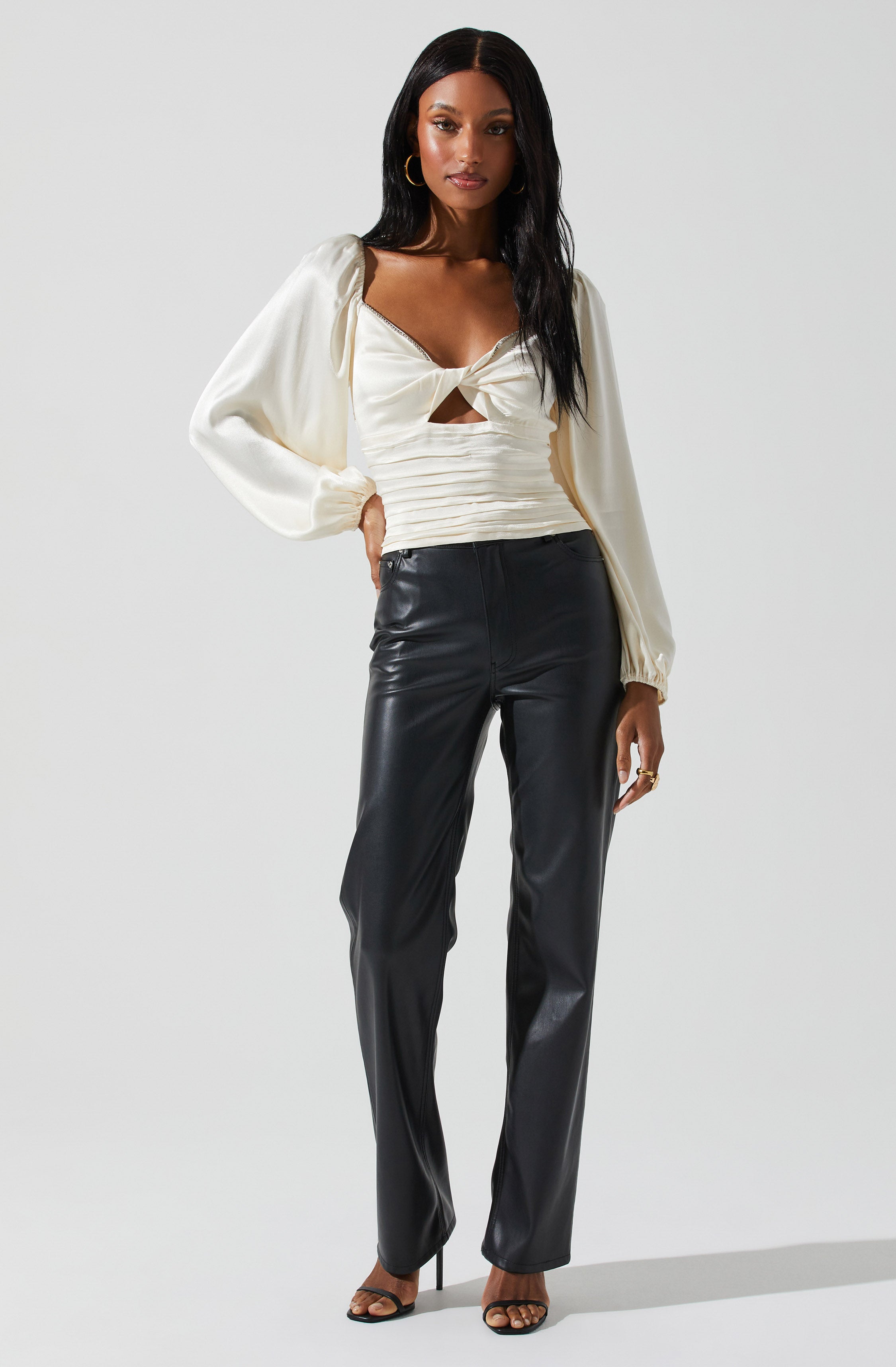 A woman is wearing a cream-colored satin blouse with long, puffed sleeves and a large knot tied at the front, revealing a small cutout. She has on high-waisted black leather pants and black high-heeled sandals. She accessorizes with hoop earrings and rings.