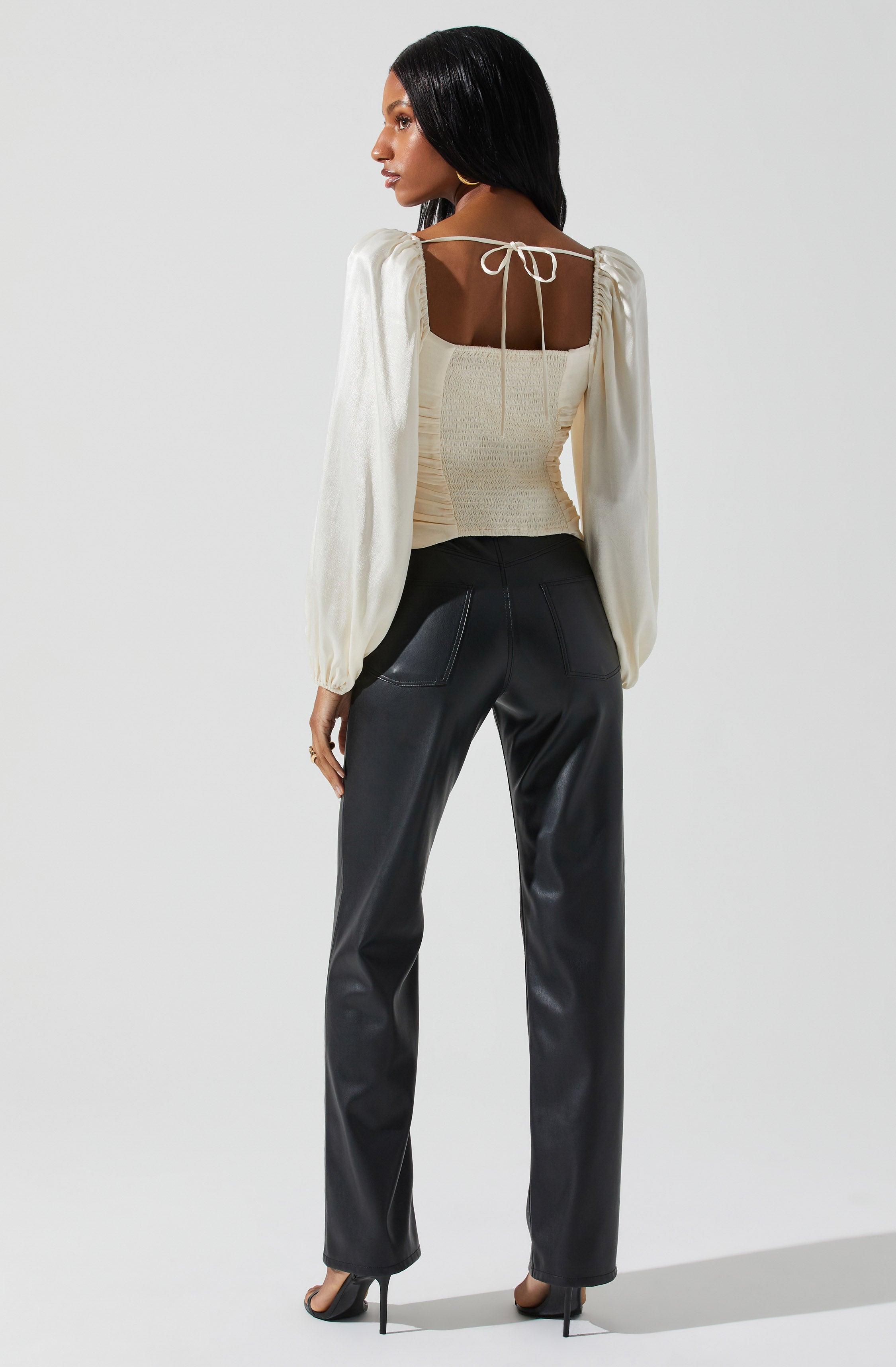 A woman is wearing a cream-colored blouse with long, puffy sleeves that are gathered at the wrists. The blouse has a square neckline with a tie string at the back. She is also wearing black leather pants with visible stitching on the pockets and a pair of black high-heeled shoes.