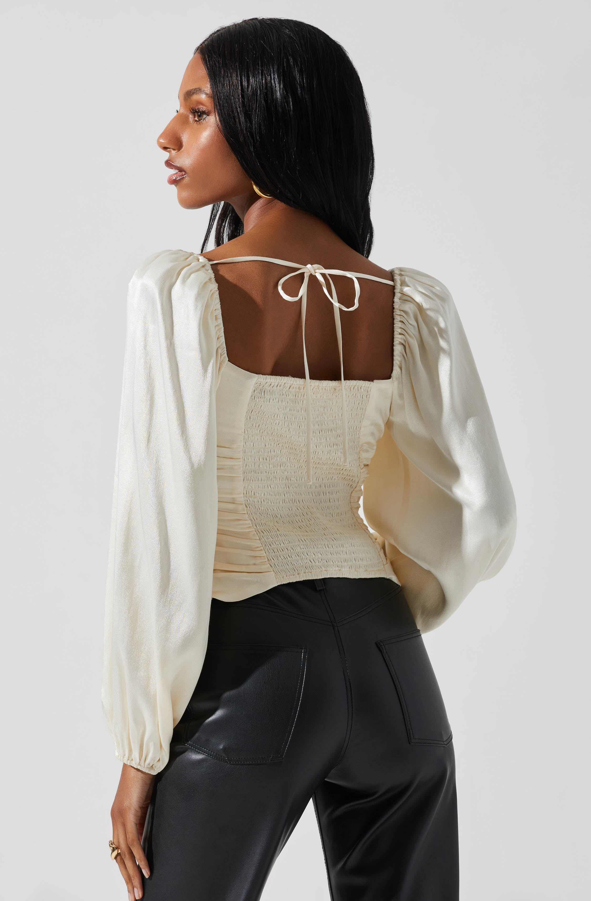 A woman wearing a cream-colored, smocked, square-neck top with puffed long sleeves and a tied string at the back. She has long, straight black hair and is also dressed in black leather pants with back patch pockets.