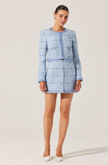 A woman is wearing a light blue and white textured two-piece outfit consisting of a cropped jacket with long sleeves and a round neckline, and a matching high-waisted skirt with front buttons. The fabric has a boucle or tweed-like appearance, giving it a classic, sophisticated look. She is also wearing white pointed-toe heels.