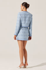 A woman is wearing a matching light blue and white tweed jacket and mini skirt set. The jacket has long sleeves and a boxy fit, while the skirt is fitted and ends above the knees. She is also wearing white high-heeled shoes.