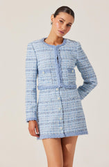 A woman is wearing a light blue, textured, tweed-style jacket and matching mini skirt set. The jacket has long sleeves, a rounded neckline, and is accented with white buttons down the front. The pattern consists of horizontal and vertical threads creating a woven appearance. The skirt sits at the waist and features similar buttons down the front. The outfit has a classic, elegant, and slightly retro style.