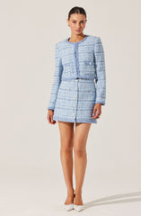 A woman is wearing a light blue and white woven tweed skirt suit. The suit includes a long-sleeved, cropped jacket with a round neckline and a matching high-waisted skirt with front buttons. The material has a textured, checkered pattern, and the ensemble is paired with white pointed-toe heels.