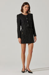 A woman is wearing a matching black tweed jacket and skirt set. The jacket has long sleeves, structured shoulders, and large decorative buttons down the front. The skirt is high-waisted, short, and features small decorative buttons on the sides. She is also wearing black pointed-toe heels with a gold embellishment on the toe and a thin ankle strap.