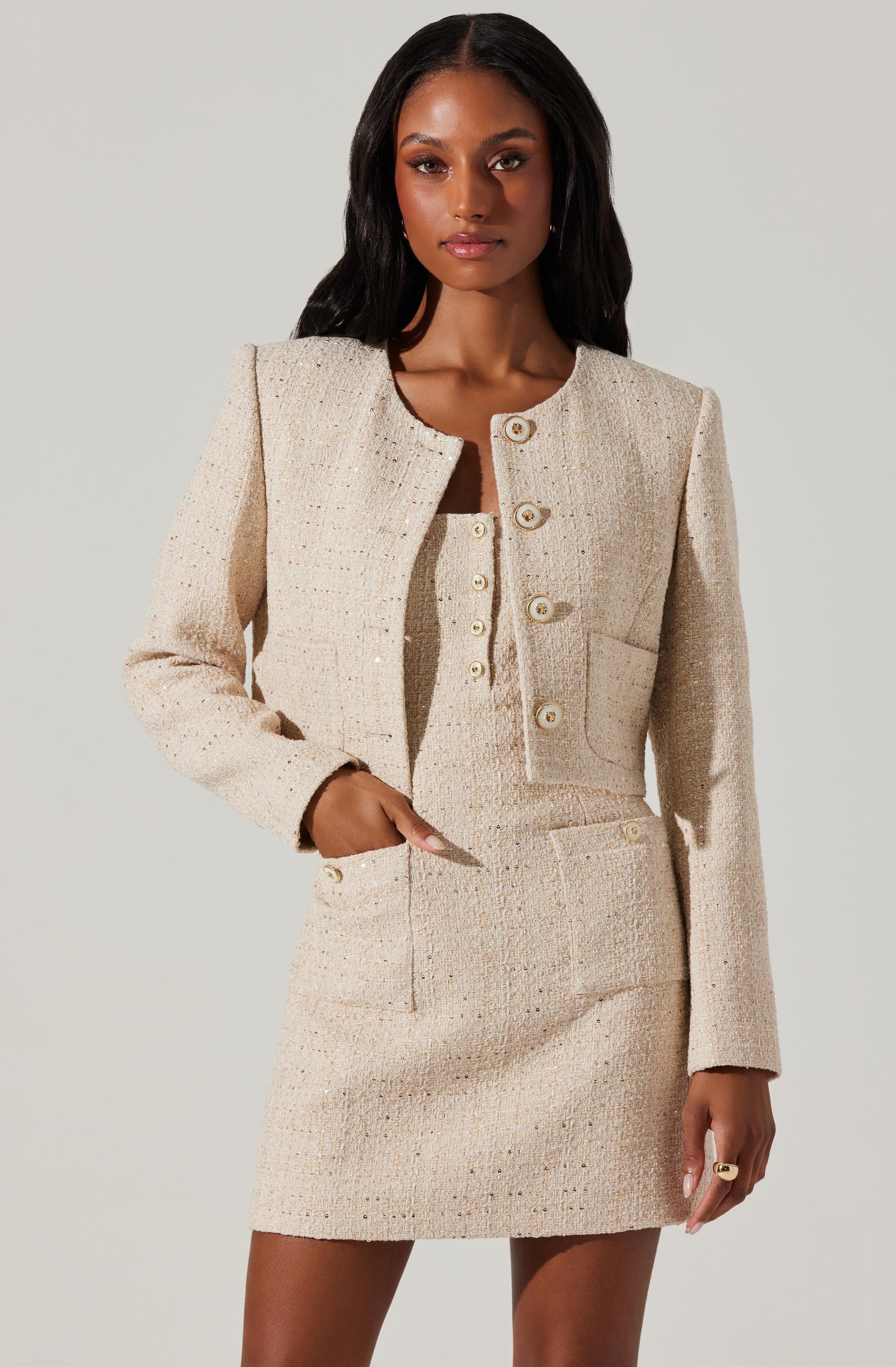 A woman is wearing a coordinated two-piece outfit in a light, cream-colored tweed fabric. The ensemble includes a fitted, high-waisted skirt that reaches mid-thigh, and a matching cropped jacket with long sleeves, a round neckline, and large buttons down the front. The jacket has a single chest pocket on the left side. The fabric has a textured appearance with small, colorful specks woven throughout.