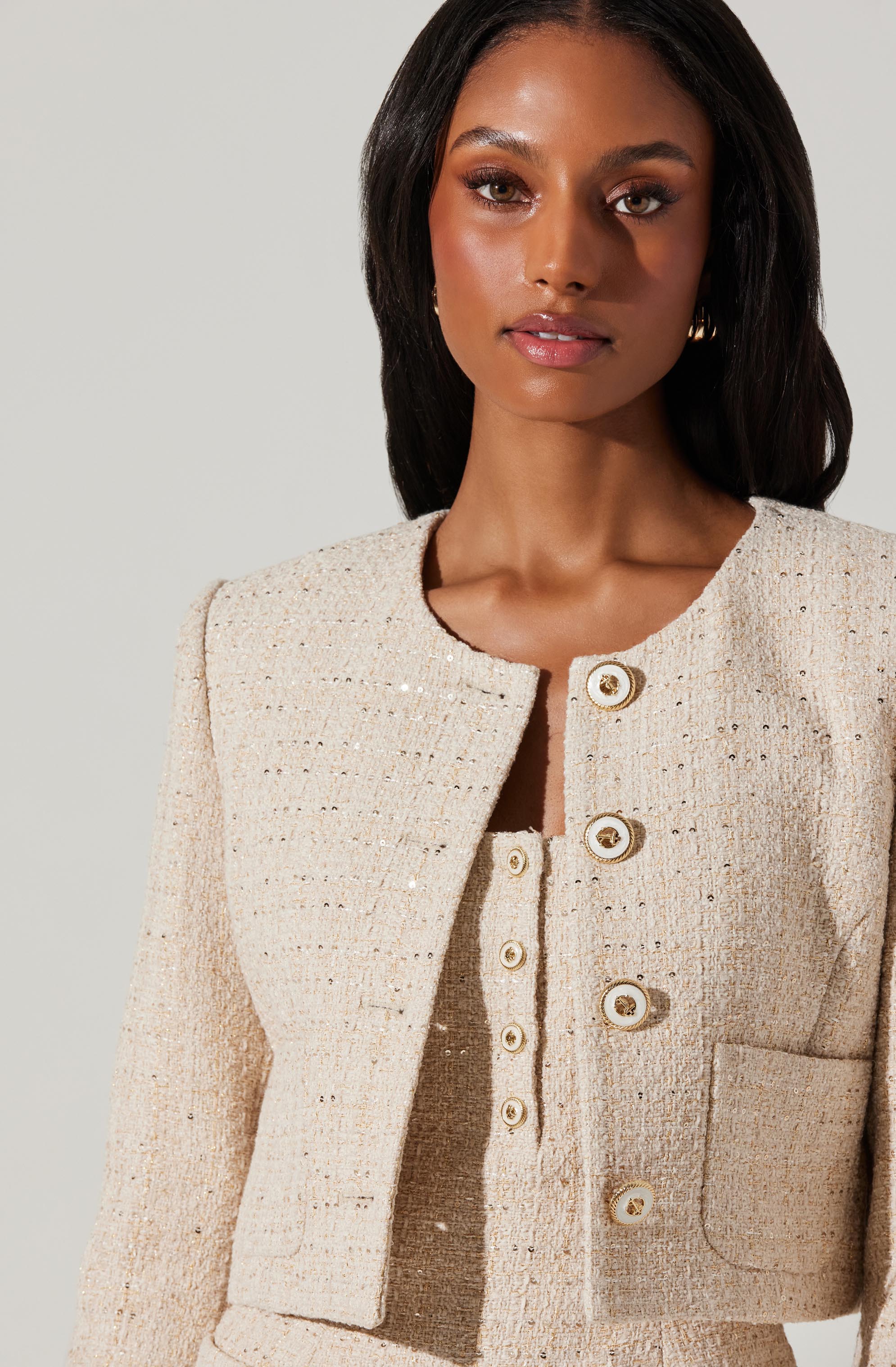 A woman is wearing a light-colored, textured, tweed-style cropped jacket with a round neckline. The jacket features a row of large, round, shiny buttons down the front and has a patch pocket on the right side. She is also wearing matching high-waisted, textured, tweed-style pants. The ensemble has a sophisticated and elegant look.