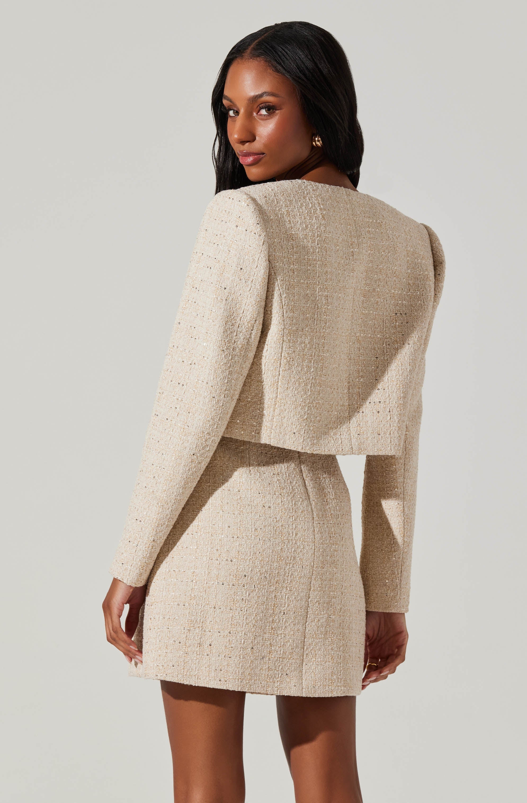 A woman wearing a matching light beige textured tweed jacket and skirt set. The jacket is cropped with long sleeves and structured shoulders. The skirt is fitted and falls above the knees.