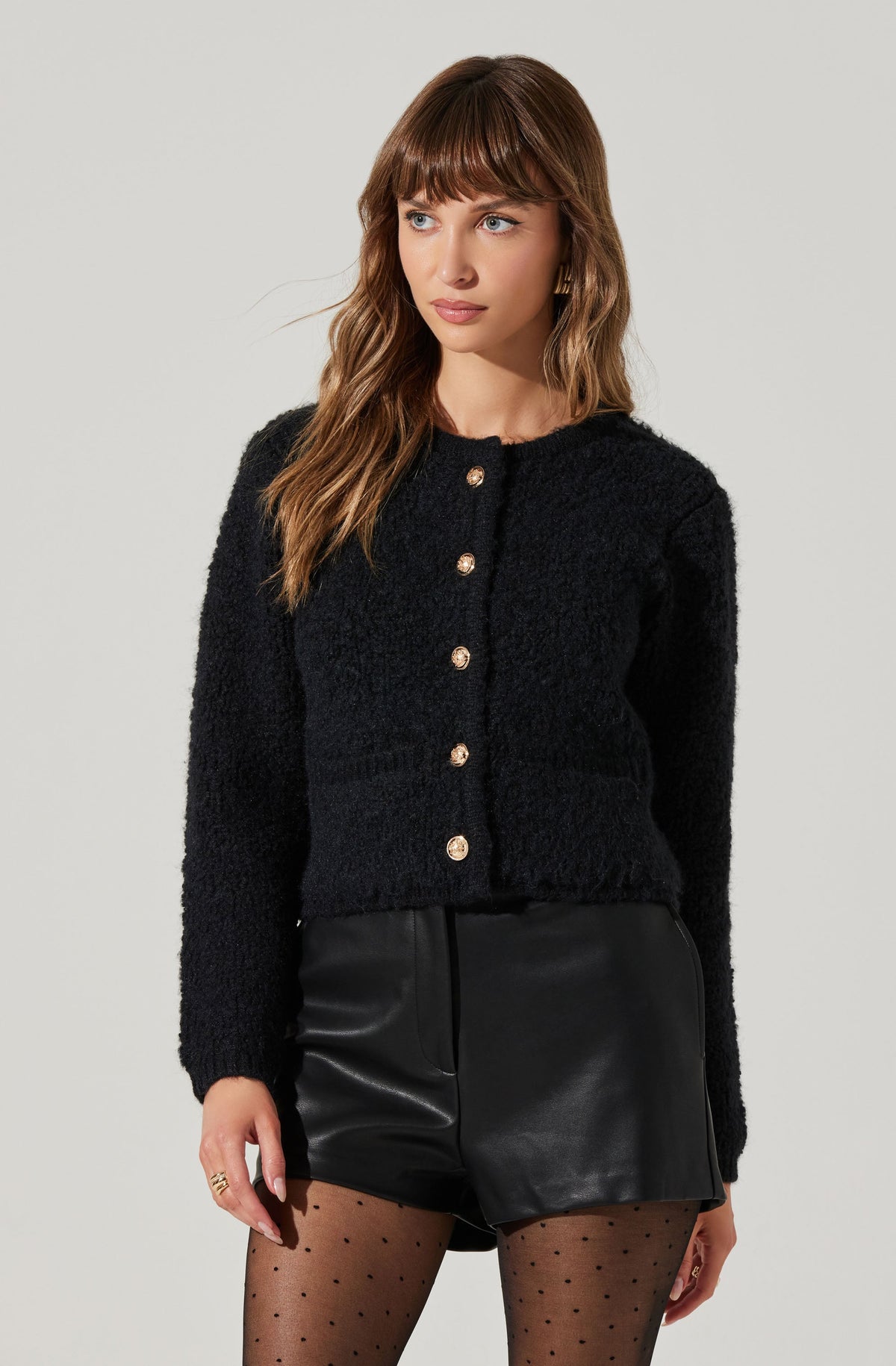 A woman is wearing a black, textured, long-sleeve button-up cardigan with gold buttons, paired with high-waisted black leather shorts. She has on black sheer tights with small polka dots.