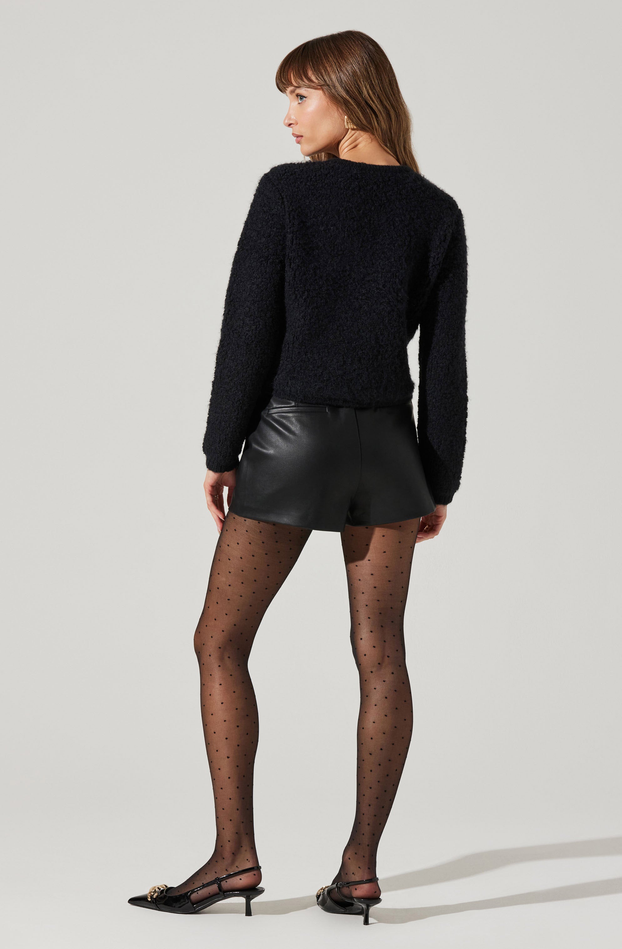 A woman is wearing a black, fuzzy long-sleeve sweater, black leather shorts, sheer black tights with small polka dots, and black high-heeled shoes with ankle straps and decorative bow details.