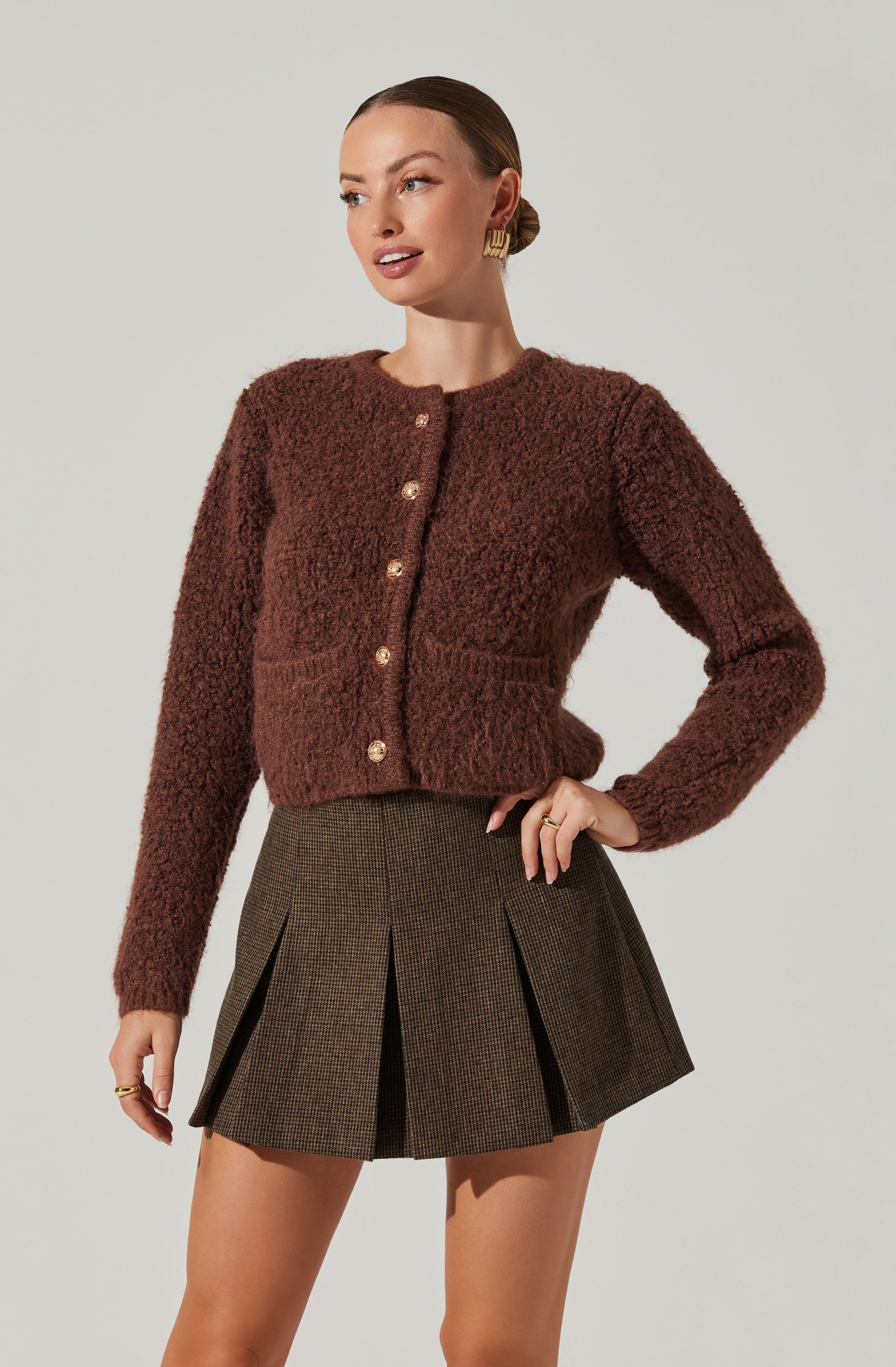 A woman is wearing a plush, textured, dark brown cardigan with large gold buttons down the front. The cardigan has a rounded neckline and long sleeves. She pairs it with a high-waisted, pleated skirt in a dark, checkered pattern. She has accessorized with gold hoop earrings and rings.