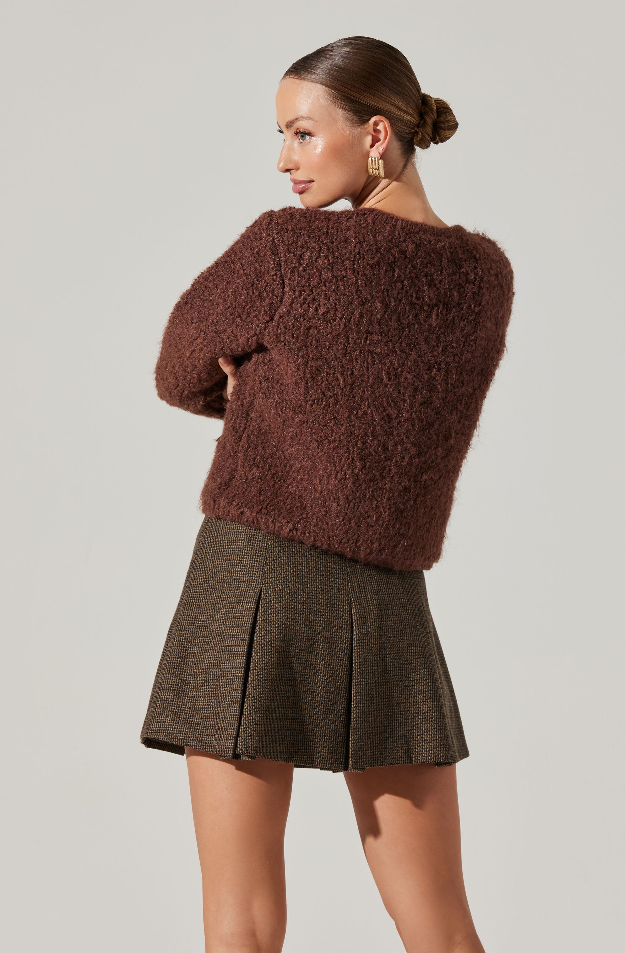 A woman is wearing a fuzzy, brown, long-sleeve sweater and a high-waisted, pleated skirt with a small checked pattern in brown and black.