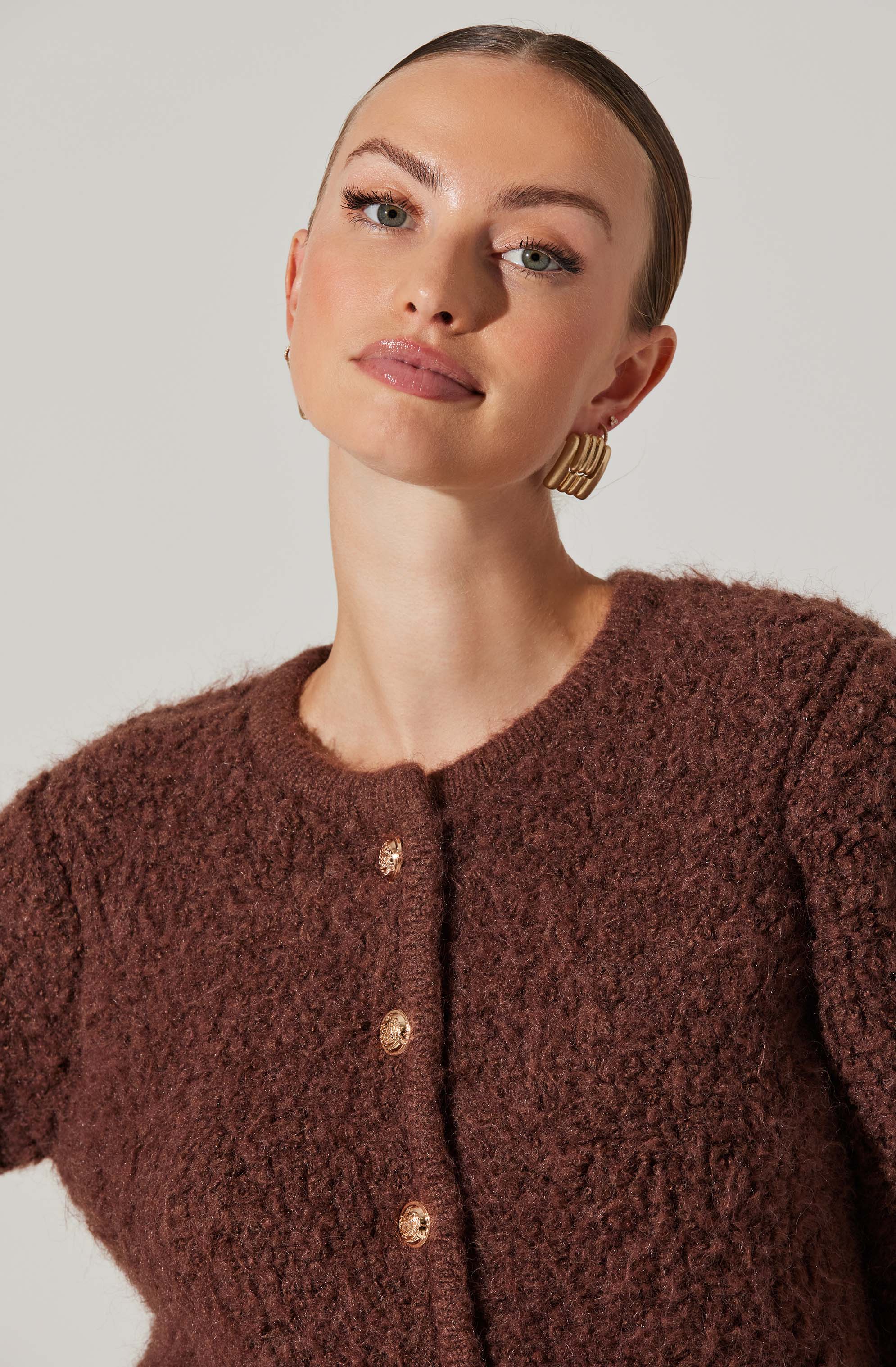 A woman wearing a textured, dark brown, button-up cardigan with a round neckline. She has her hair slicked back and is accessorized with large, gold, rectangular earrings.