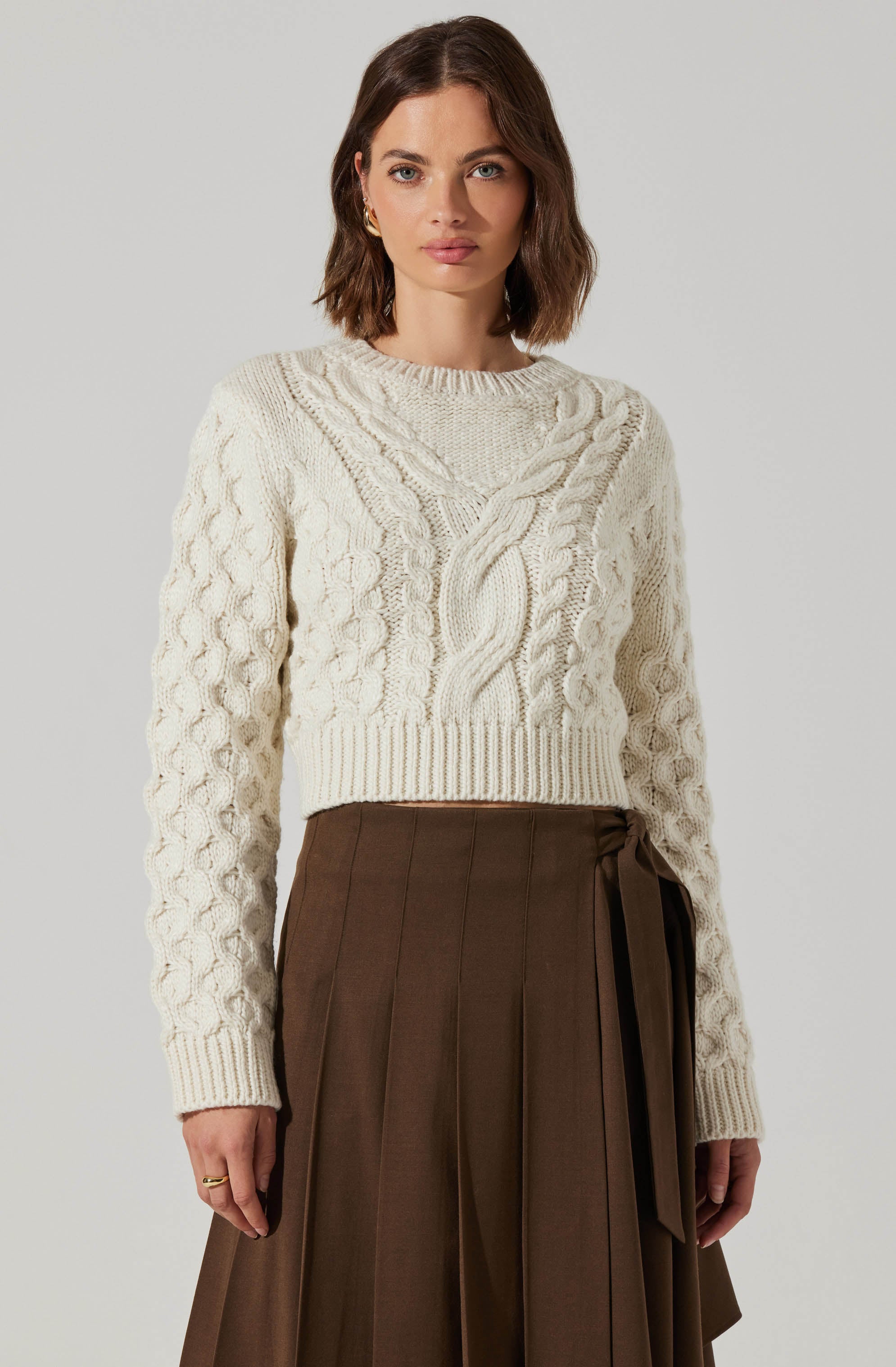 A woman is wearing a cream-colored, cable knit sweater with a variety of braided and textured knit patterns, including a prominent central braid and smaller braided details throughout. The sweater has a crew neckline and long sleeves. She pairs it with a high-waisted, dark brown pleated skirt that features a tied sash detail on one side.