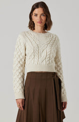 A woman is wearing a cream-colored, cable knit sweater with a variety of braided and textured knit patterns, including a prominent central braid and smaller braided details throughout. The sweater has a crew neckline and long sleeves. She pairs it with a high-waisted, dark brown pleated skirt that features a tied sash detail on one side.