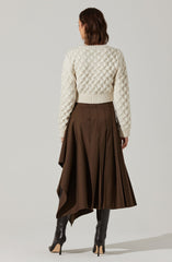 A woman is wearing a cream-colored knit sweater with a textured, cable-knit pattern, a wide ribbed hem, and long sleeves. She pairs it with a high-waisted, brown pleated skirt that falls below the knees, along with black high-heeled boots.