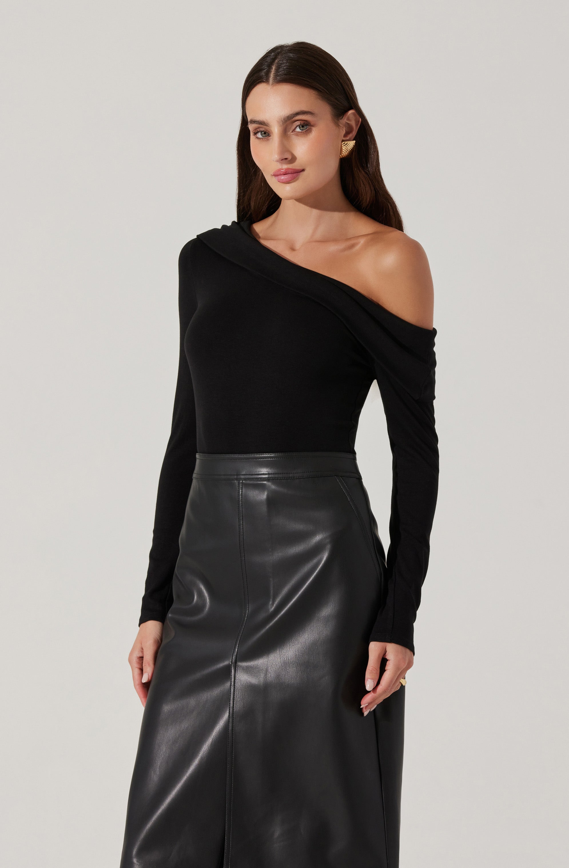 A woman is wearing a black off-the-shoulder top with long sleeves and a draped neckline. She pairs it with a high-waisted, black leather skirt that reaches below her knees. She accessorizes with gold statement earrings and has long, dark brown hair.