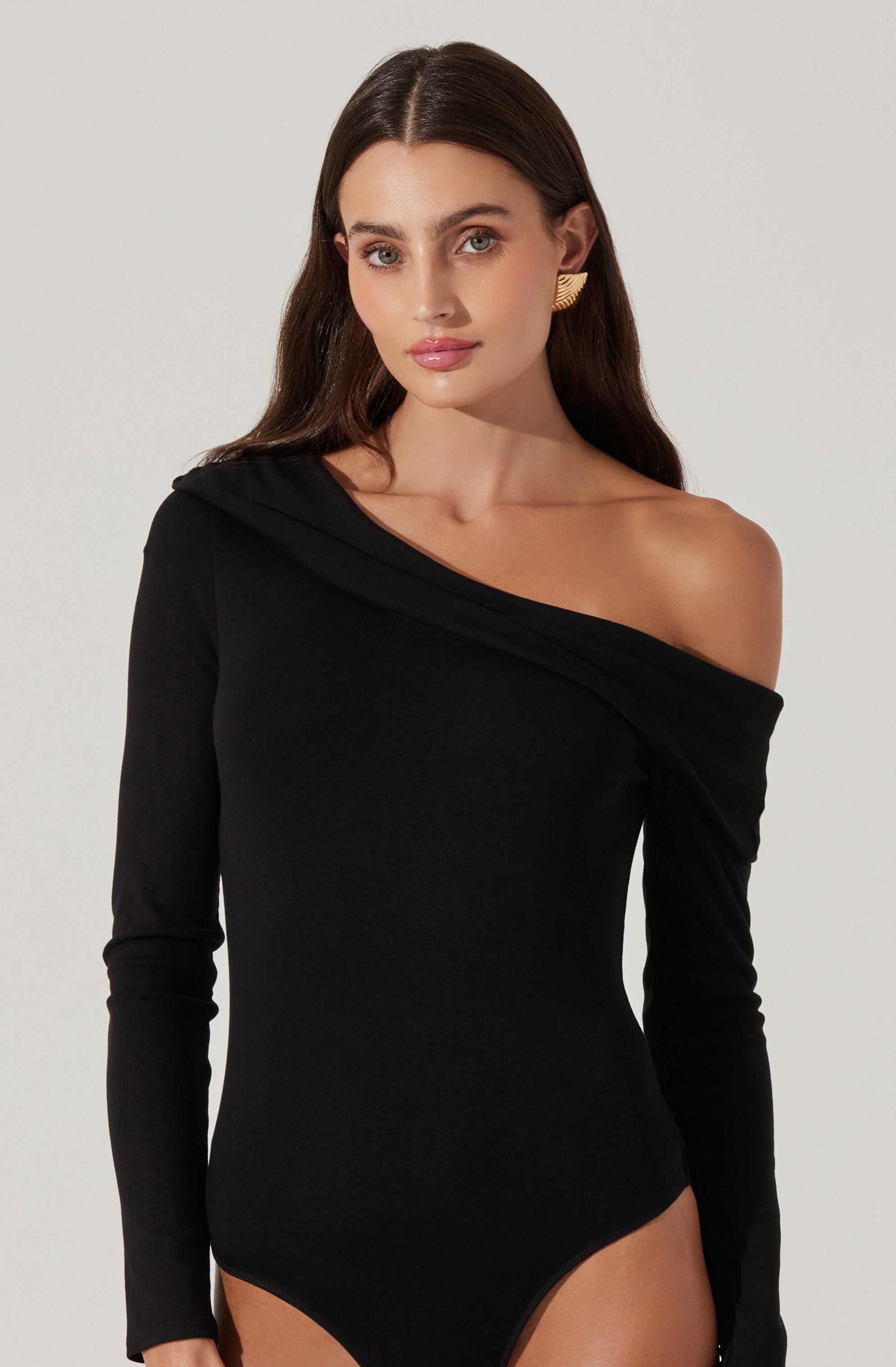 A woman wearing a black, long-sleeved bodysuit with an asymmetrical neckline that drapes off her left shoulder. The bodysuit fits closely to her body, accentuating her figure. She is accessorized with large, gold, geometric earrings.