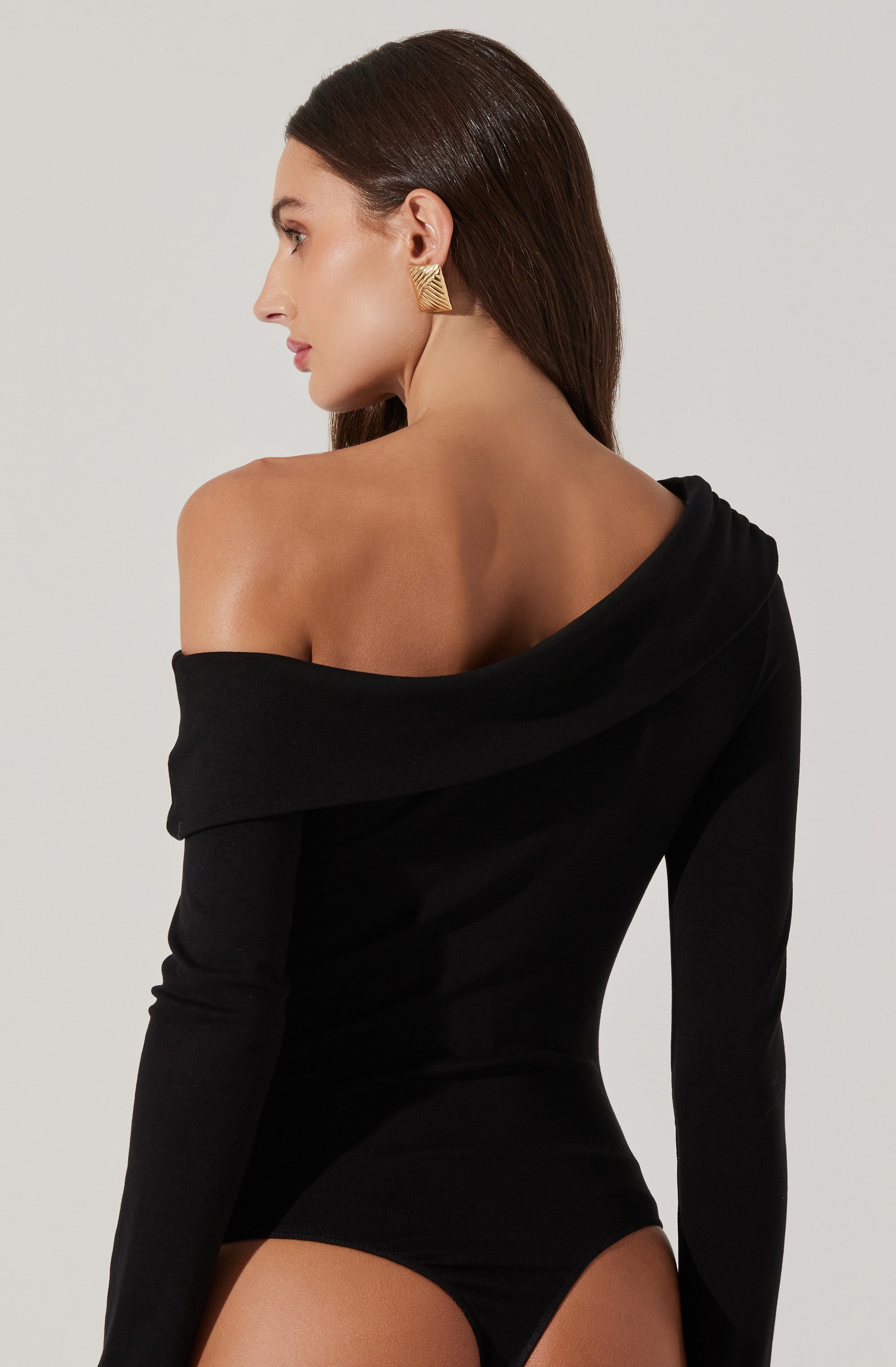 A woman is wearing a fitted black off-the-shoulder long-sleeve bodysuit. The garment has a wide neckline that drapes over one shoulder, exposing her collarbone and shoulder. She accessorizes with large gold geometric earrings.