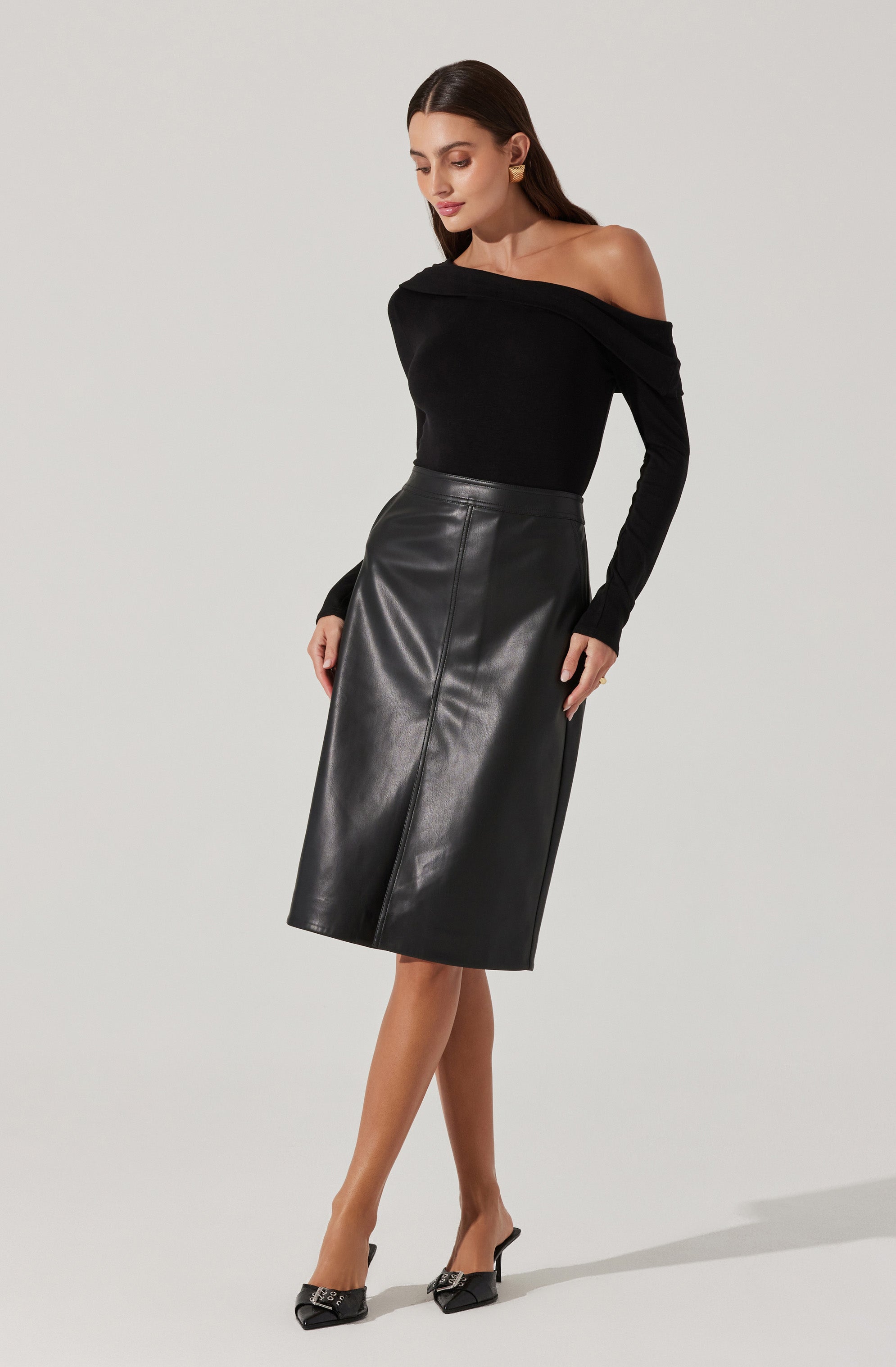 A woman is wearing an off-the-shoulder black long-sleeve top and a high-waisted, knee-length black leather skirt. She completes the look with pointed black heels that have decorative embellishments on the front.