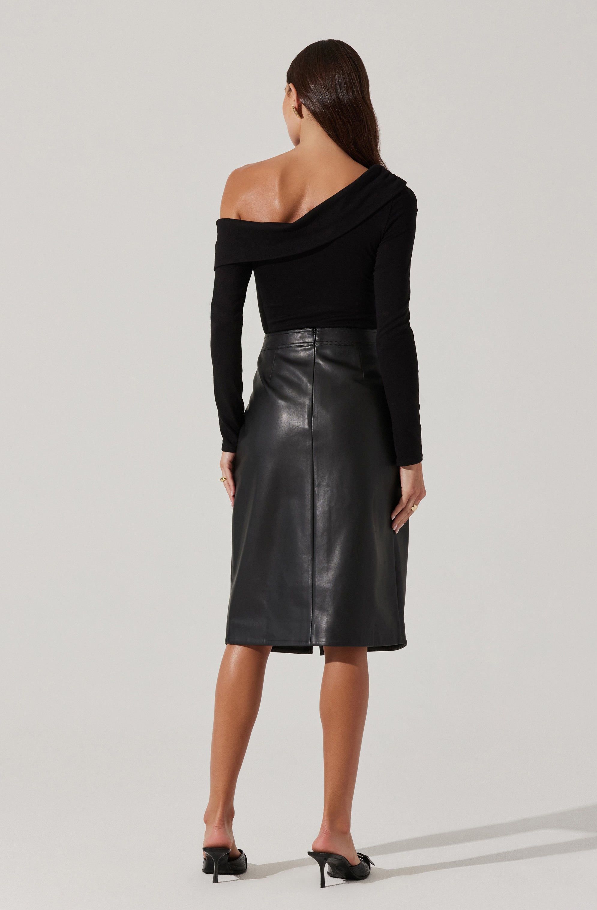 A woman is wearing a black off-the-shoulder top with long sleeves, which drapes over one shoulder, exposing her left shoulder and upper back. She pairs it with a high-waisted, black leather midi skirt that reaches below her knees. She completes her outfit with black high-heeled sandals.