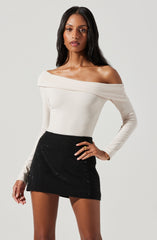 A woman is wearing an off-the-shoulder, long-sleeved white top with a wide neckline that drapes over her shoulders. She pairs it with a high-waisted, black mini skirt that has a shiny, textured surface.