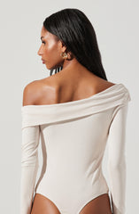 A woman is wearing an off-the-shoulder, long-sleeve bodysuit in a light cream color. The fabric appears smooth and fitted, with a draped collar that folds over the shoulders, creating a stylish and elegant look. She has gold hoop earrings and her long, straight black hair falls over her back.