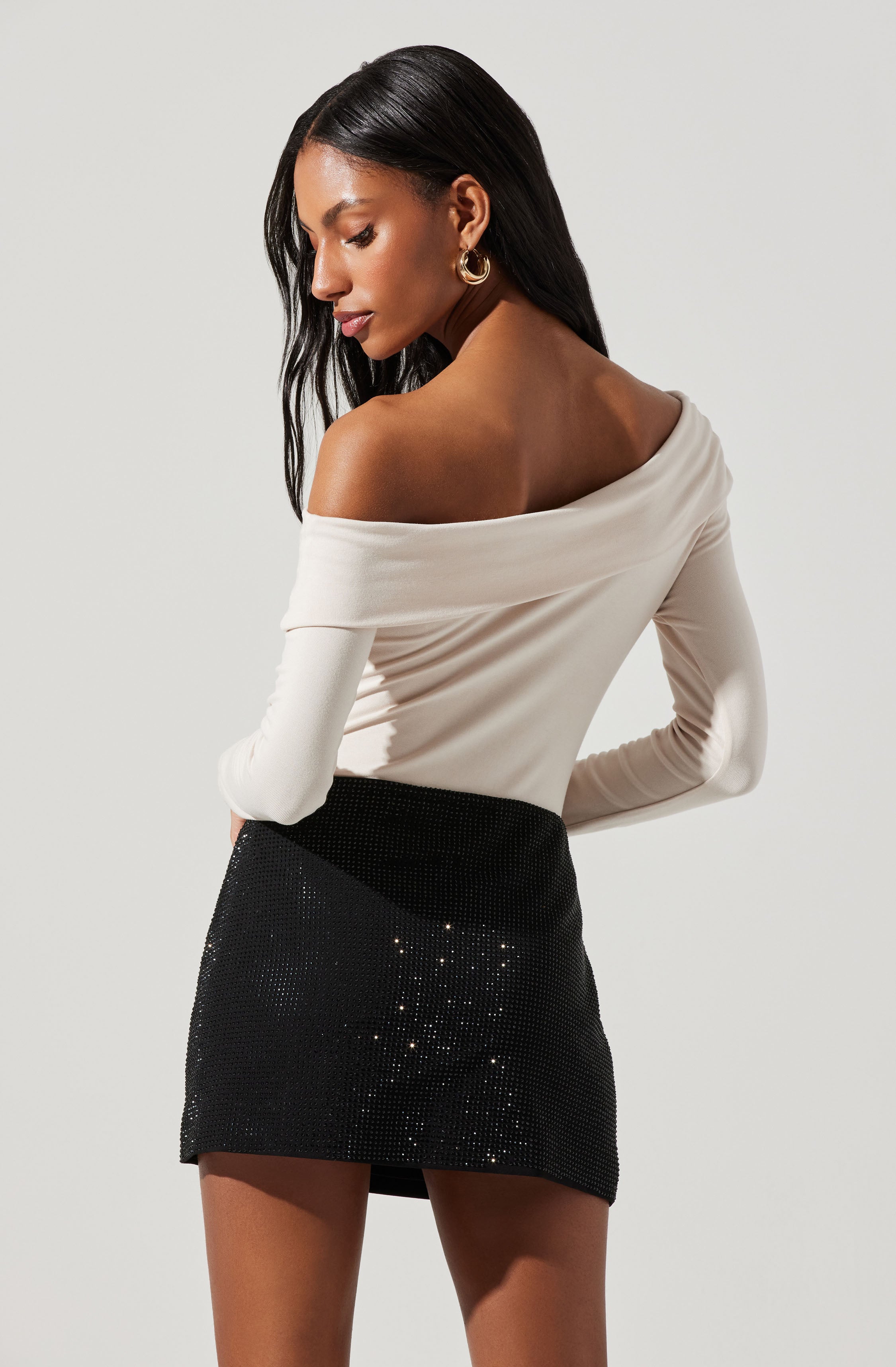 A woman is wearing an off-the-shoulder, long-sleeved, cream-colored top paired with a high-waisted black mini skirt that has a subtle shimmer or sequined embellishments.