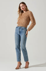 A woman is wearing a beige, fuzzy, long-sleeved sweater with a textured, slightly shaggy appearance. She pairs it with high-waisted, light blue jeans that have a classic fit and slightly cropped hem. She completes the outfit with beige high-heeled shoes featuring a pointed toe and a textured pattern. She accessorizes with gold hoop earrings and a gold ring.